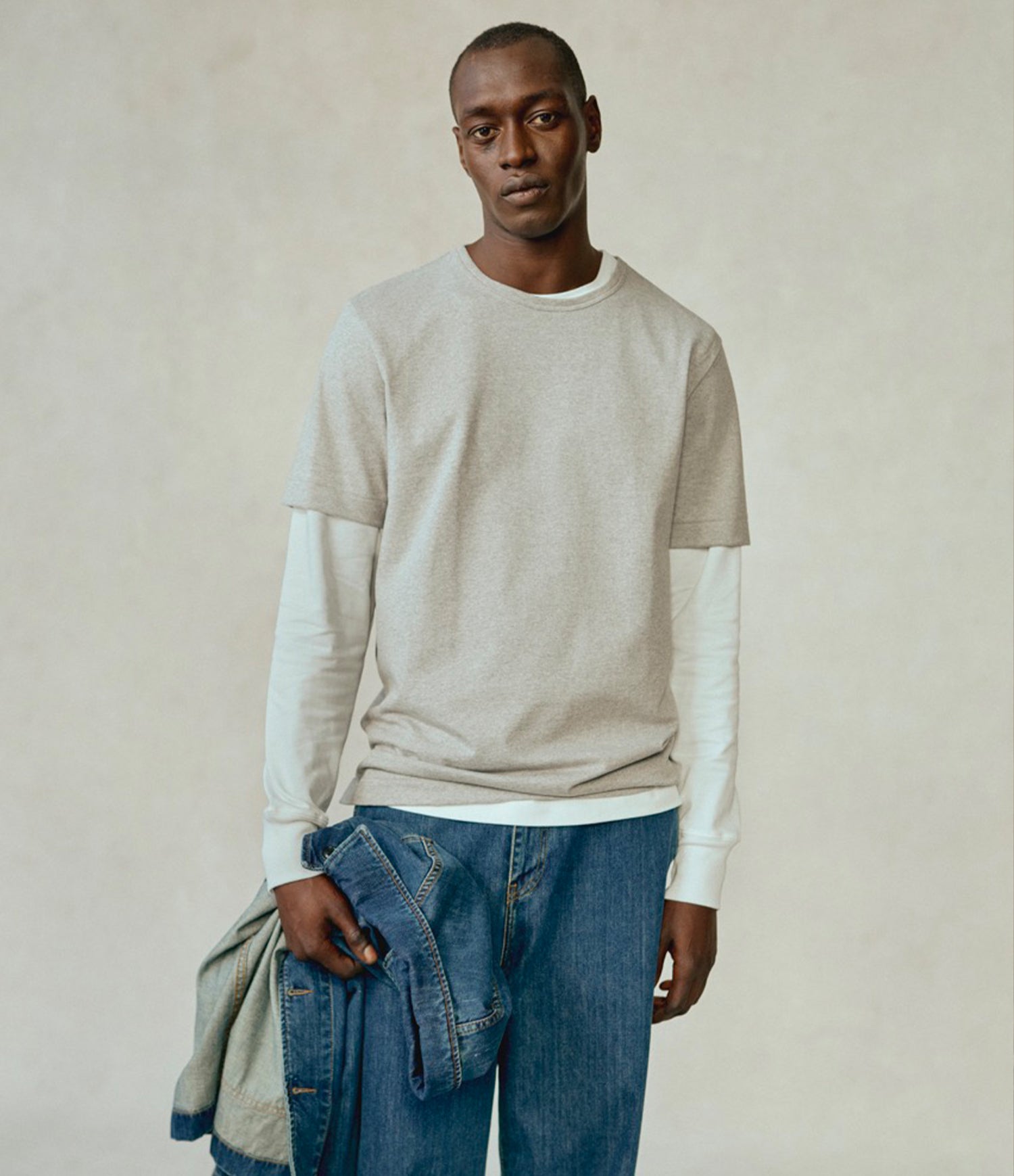 A man modeling a light beige crewneck t-shirt layered over a white long-sleeve shirt, paired with blue jeans, standing against a neutral beige textured background.