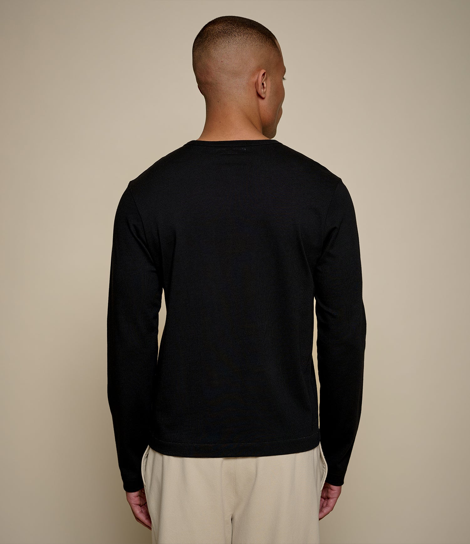 A man viewed from behind wearing a plain black long-sleeve crewneck sweater and light beige trousers, standing against a solid beige studio background.