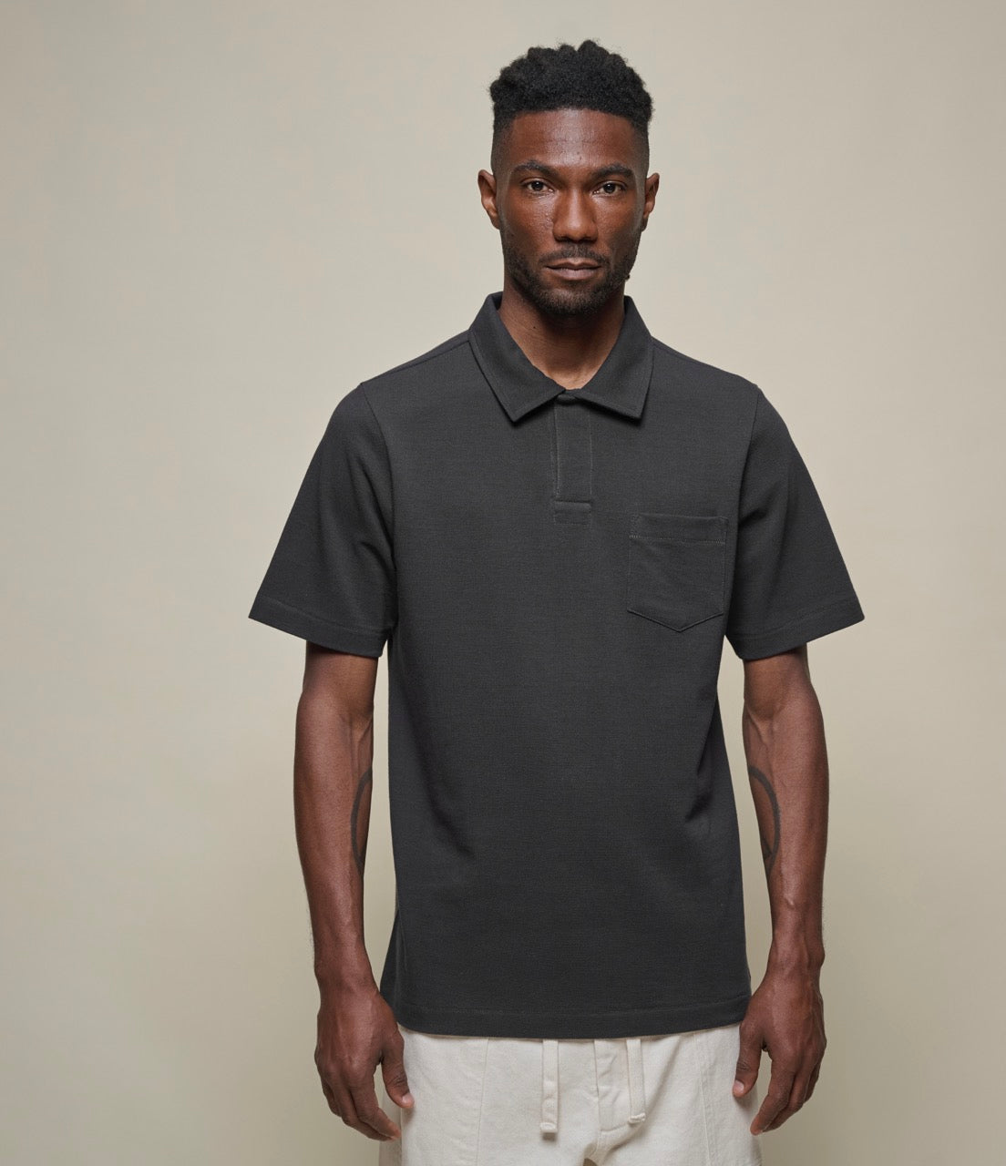 A man wearing a black short sleeve polo shirt with a collar and a front pocket, paired with light-colored shorts.