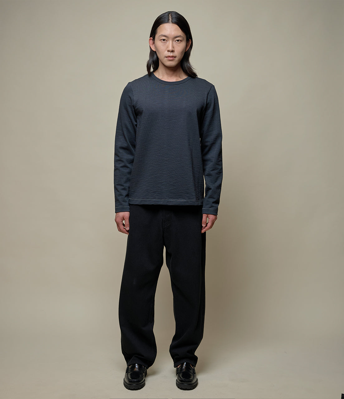 Person wearing a dark gray long-sleeve shirt and black pants against a beige background