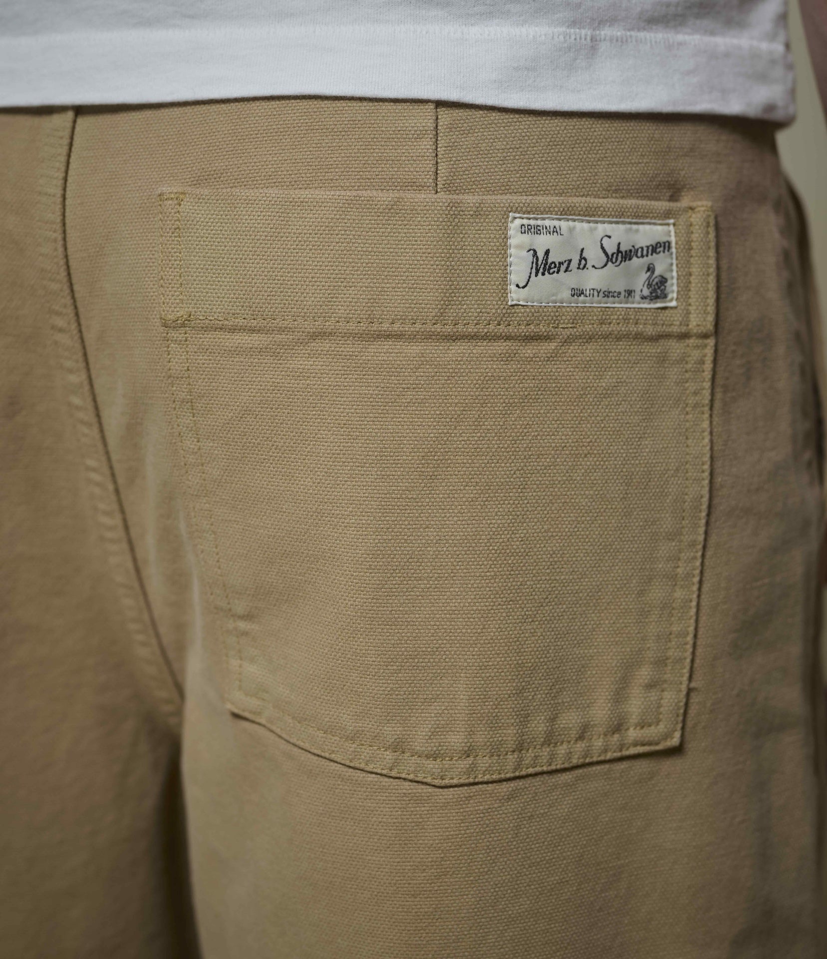 Close-up of the back pocket of tan chinos, showing a white woven label that reads 'Merz b. Schwanen', worn with a white t-shirt.