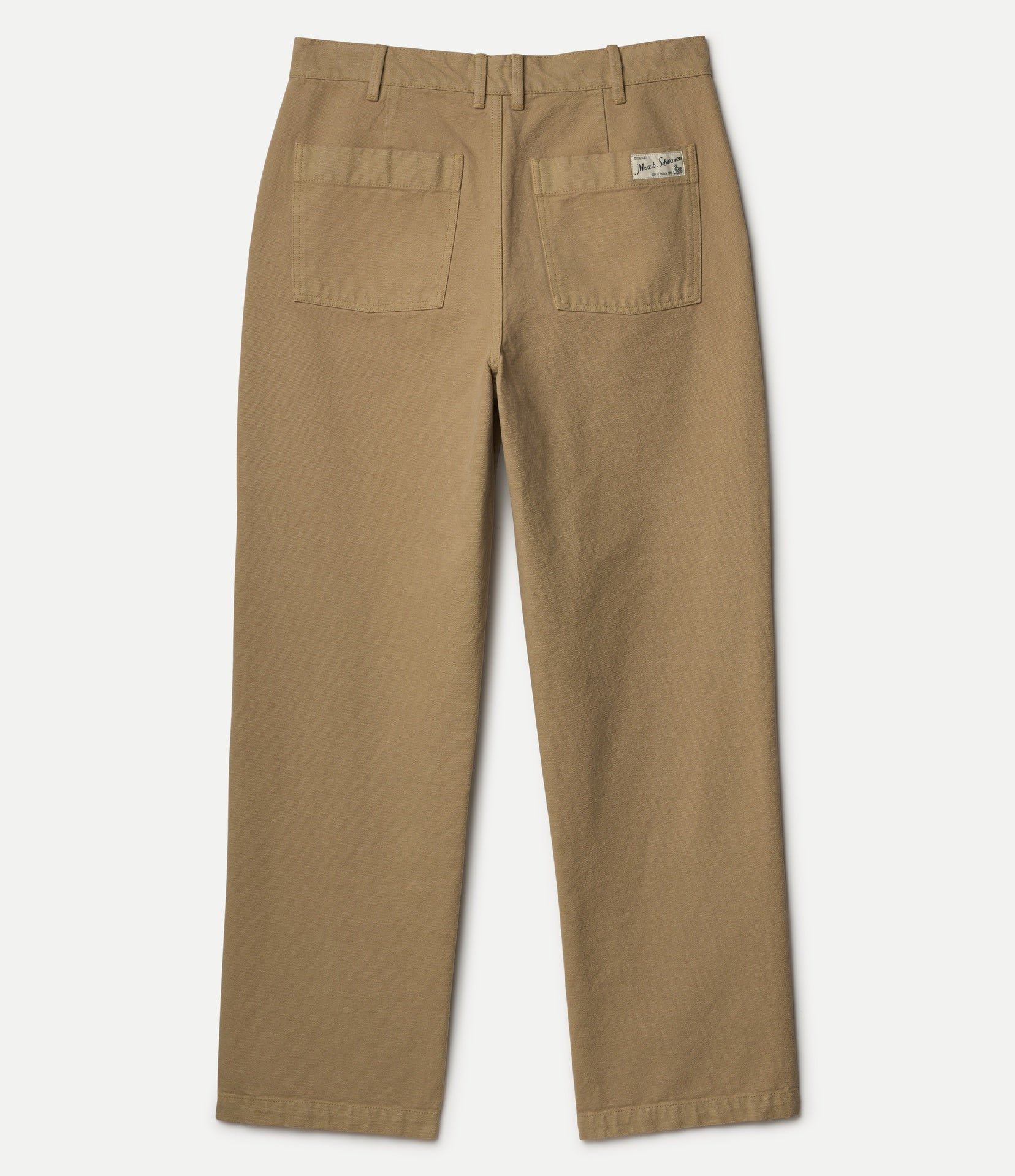 A pair of tan work pants shown from the back, featuring two large patch pockets and a small Merz b. Schwanen brand label on the right pocket, set against a plain light gray background.