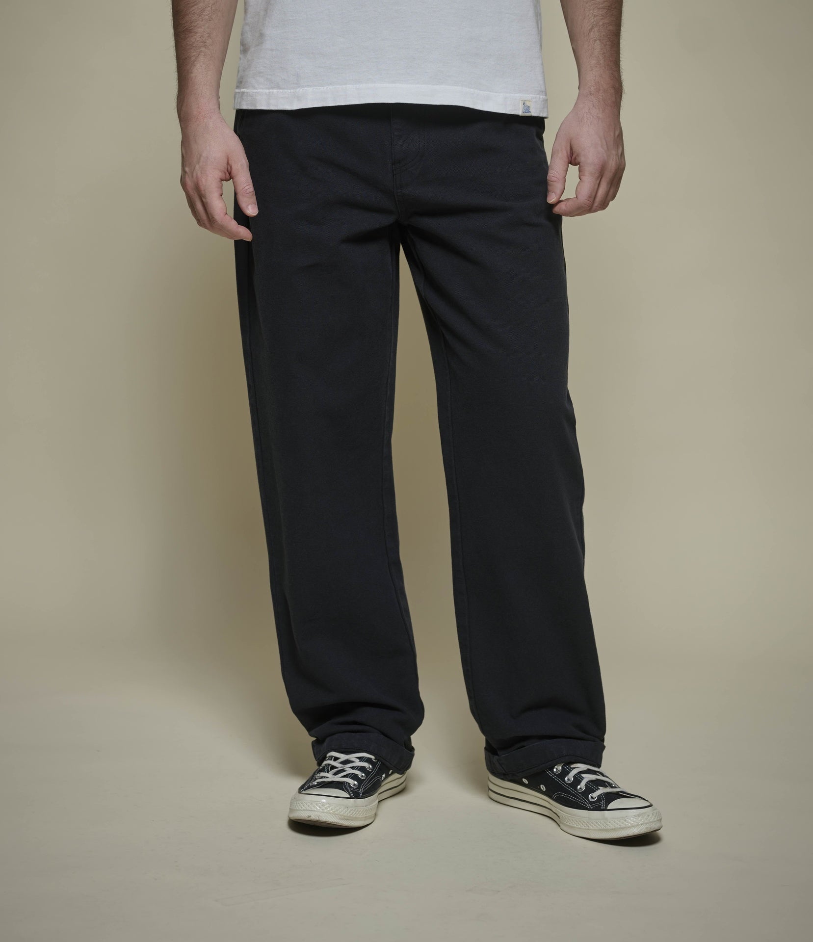 A person wearing black straight-leg pants, a white t-shirt, and black canvas sneakers, standing against a solid beige studio background.