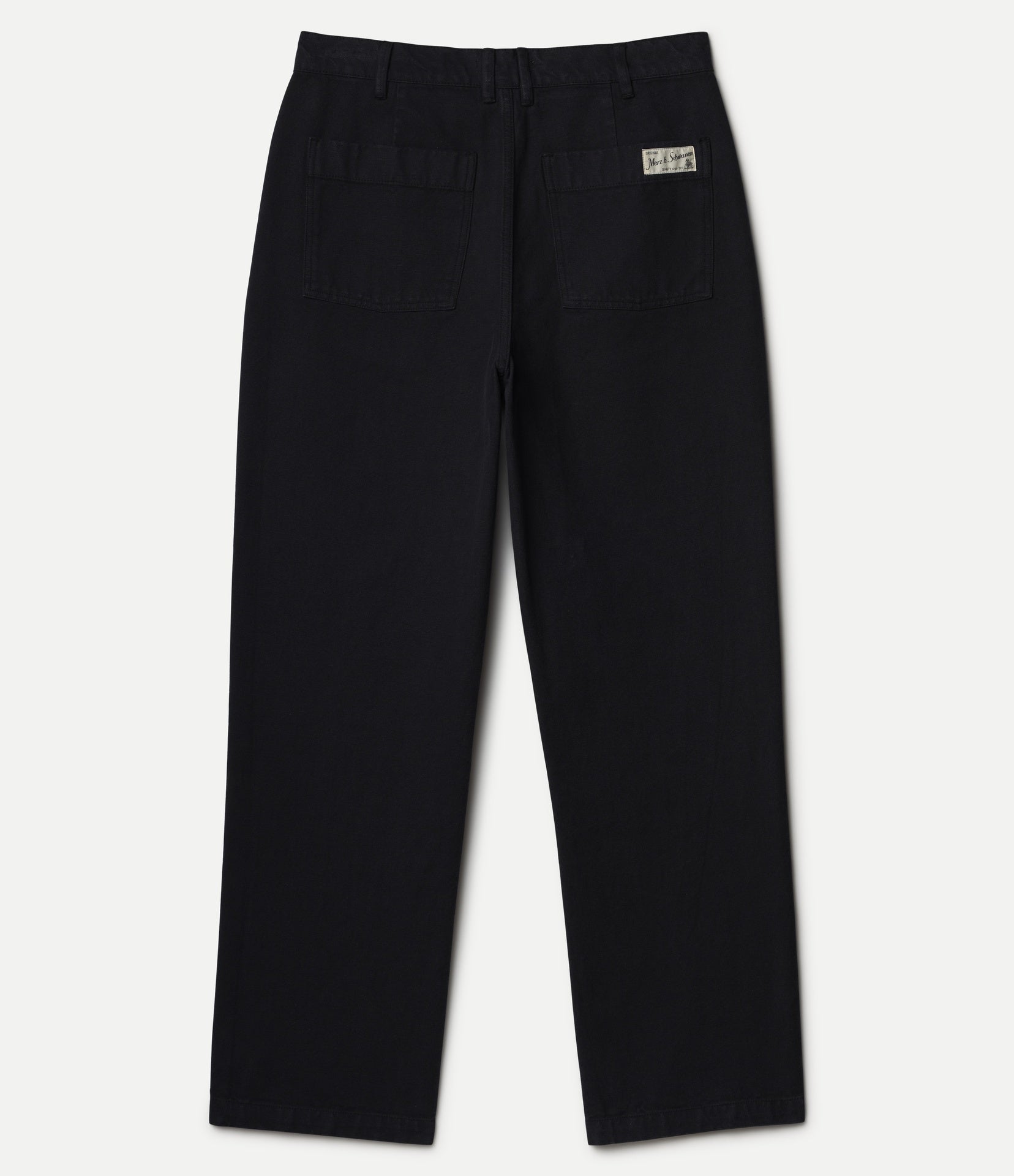 Black straight-leg pants shown from the back, featuring a button waistband, belt loops, two back patch pockets, and a small rectangular Merz b. Schwanen brand patch on the upper right side, set against a plain light background.