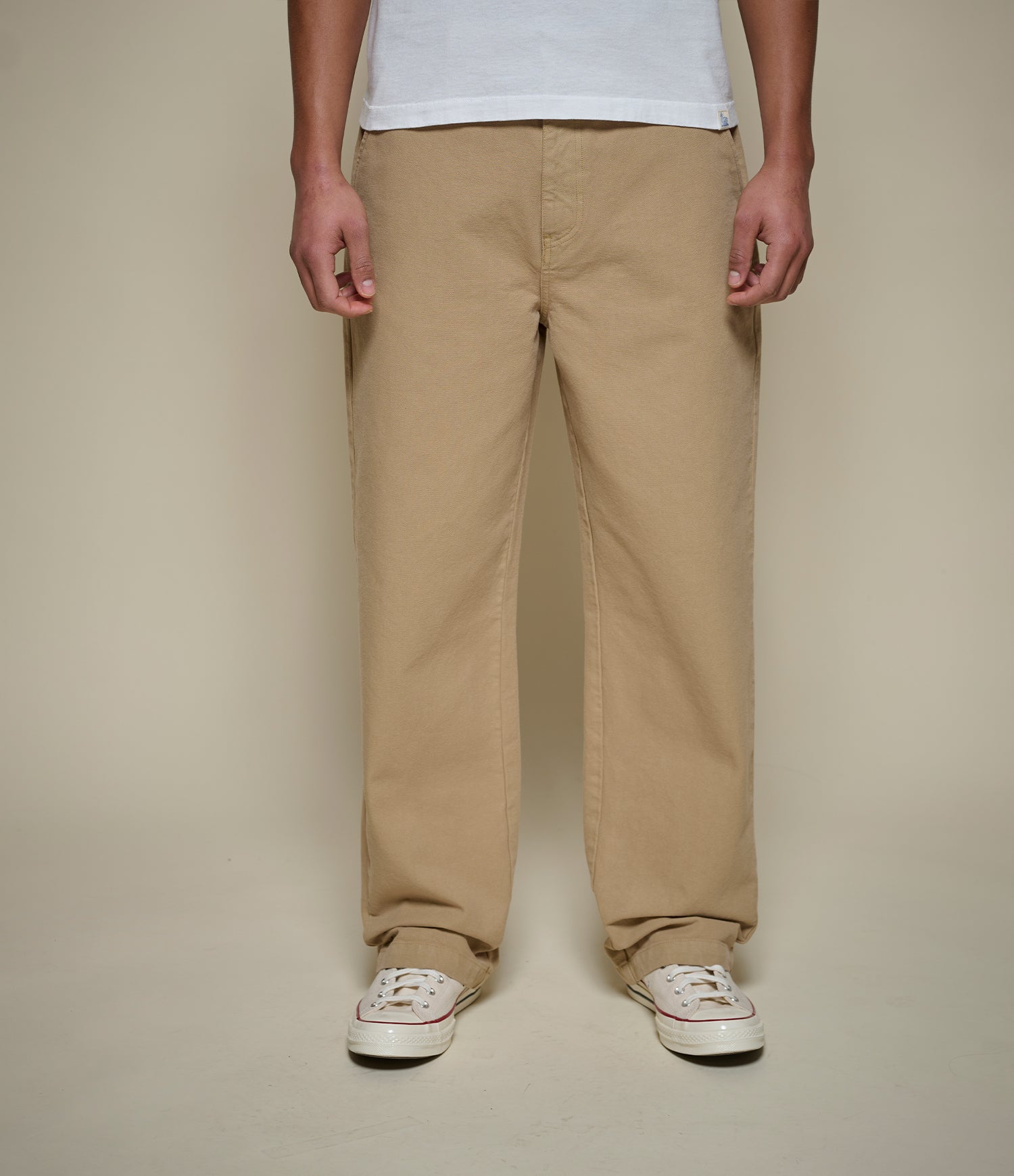A person wearing straight-leg khaki pants, a white t-shirt, and white sneakers, standing against a solid beige background.