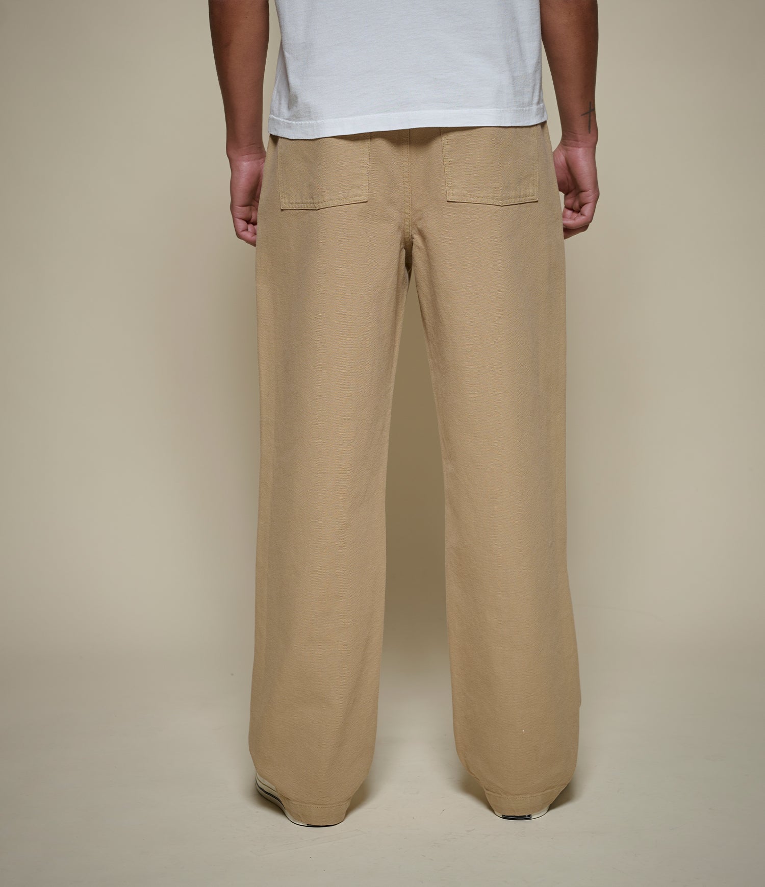 A person shown from the back wearing beige straight-leg trousers and a white t-shirt, standing against a plain beige studio background.
