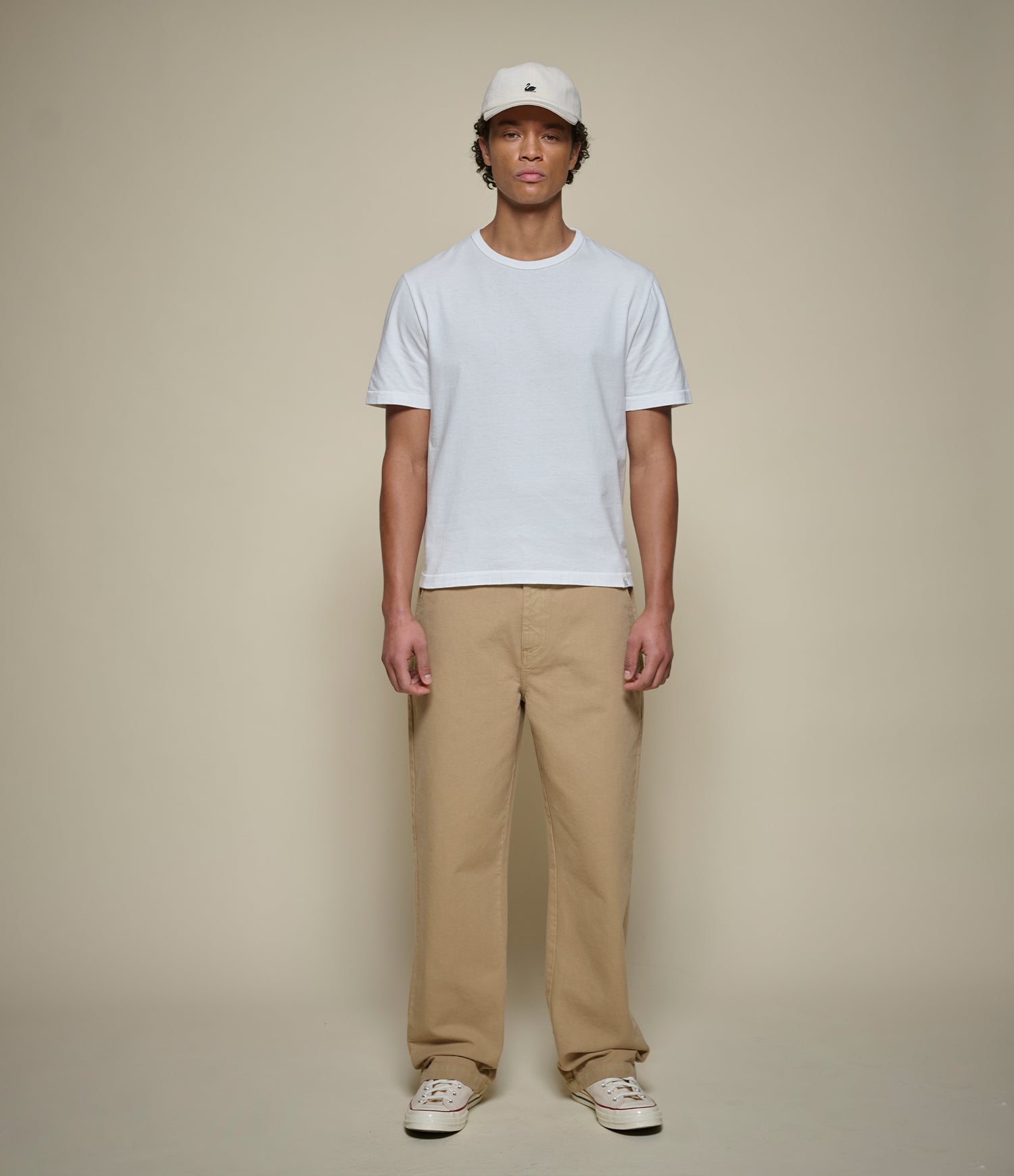 A man stands against a plain beige studio background wearing a white short-sleeve crewneck t-shirt, beige straight leg pants, white sneakers, and a white baseball cap.