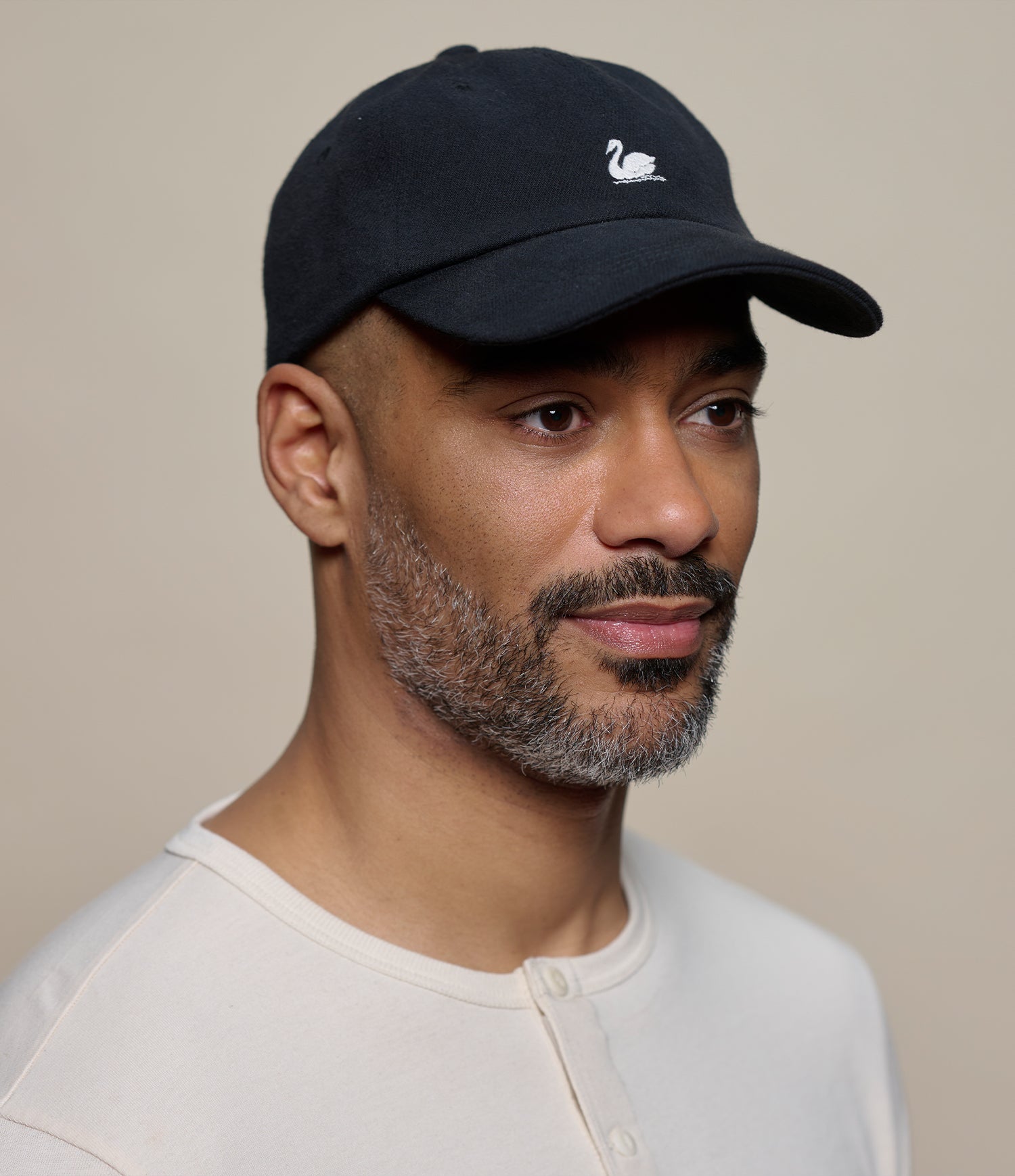 A man wearing a black baseball cap with a small white swan logo on the front, paired with a light beige henley shirt, set against a plain beige background.