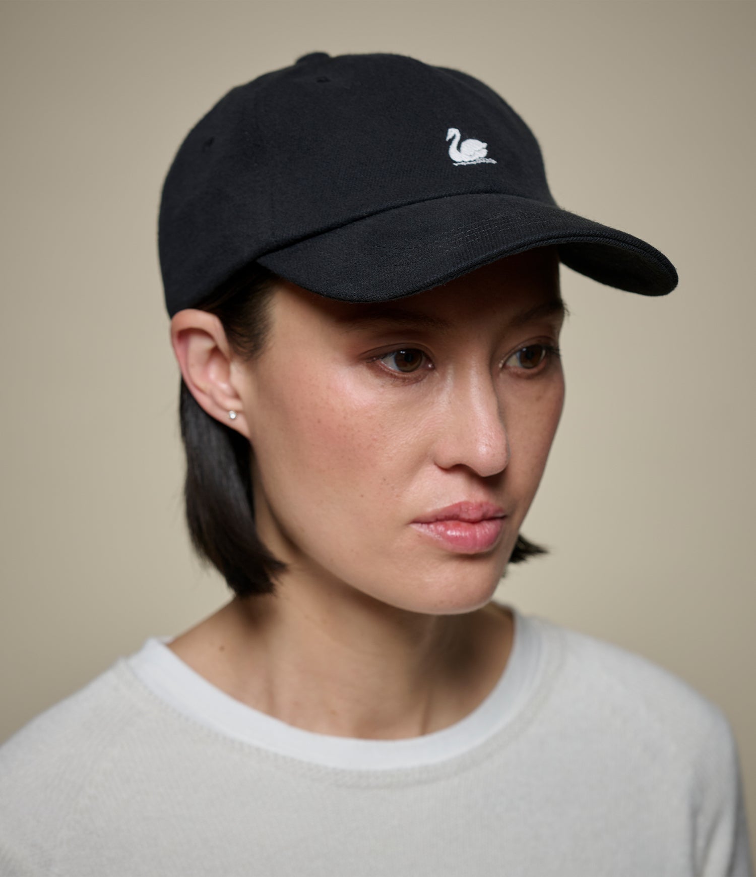 A woman wearing a black baseball cap with a small white swan logo on the front, paired with a light gray crewneck sweater, set against a plain beige background.