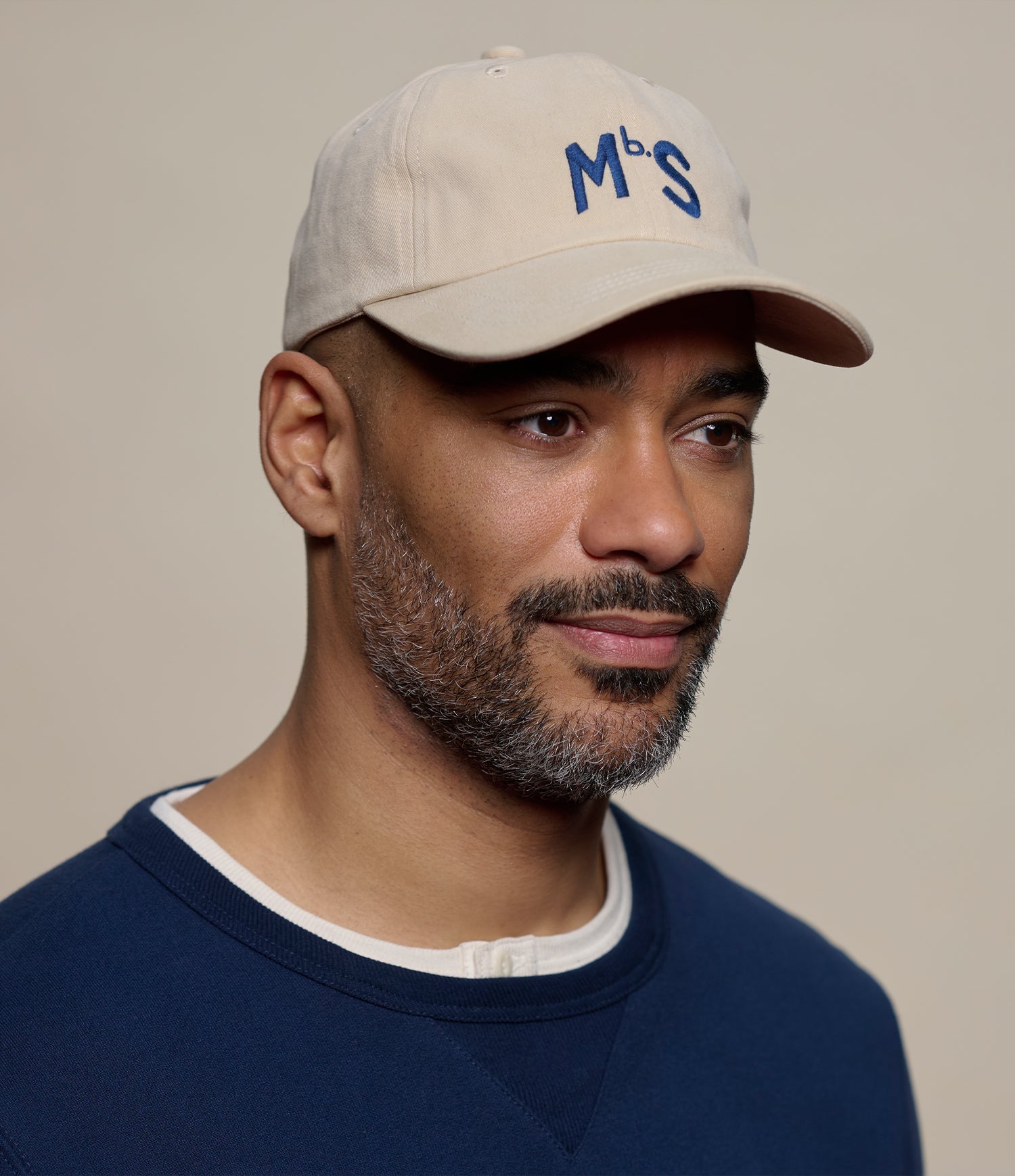 Man wearing a beige cap with 'MbS' logo and navy sweater on a plain background