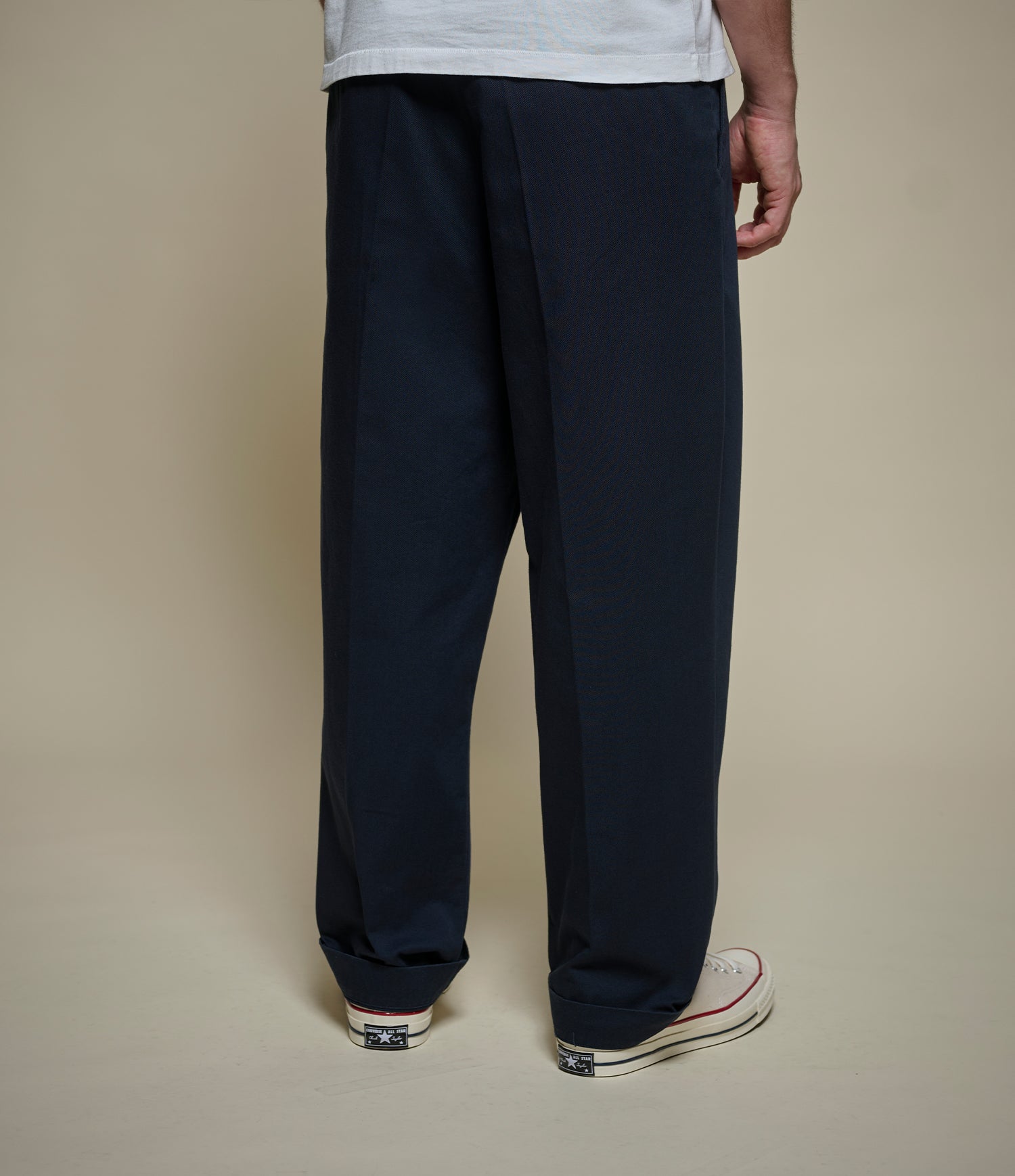 A person shown from the back wearing dark navy wide-leg trousers, a light gray t-shirt, and white canvas sneakers against a solid beige studio background.