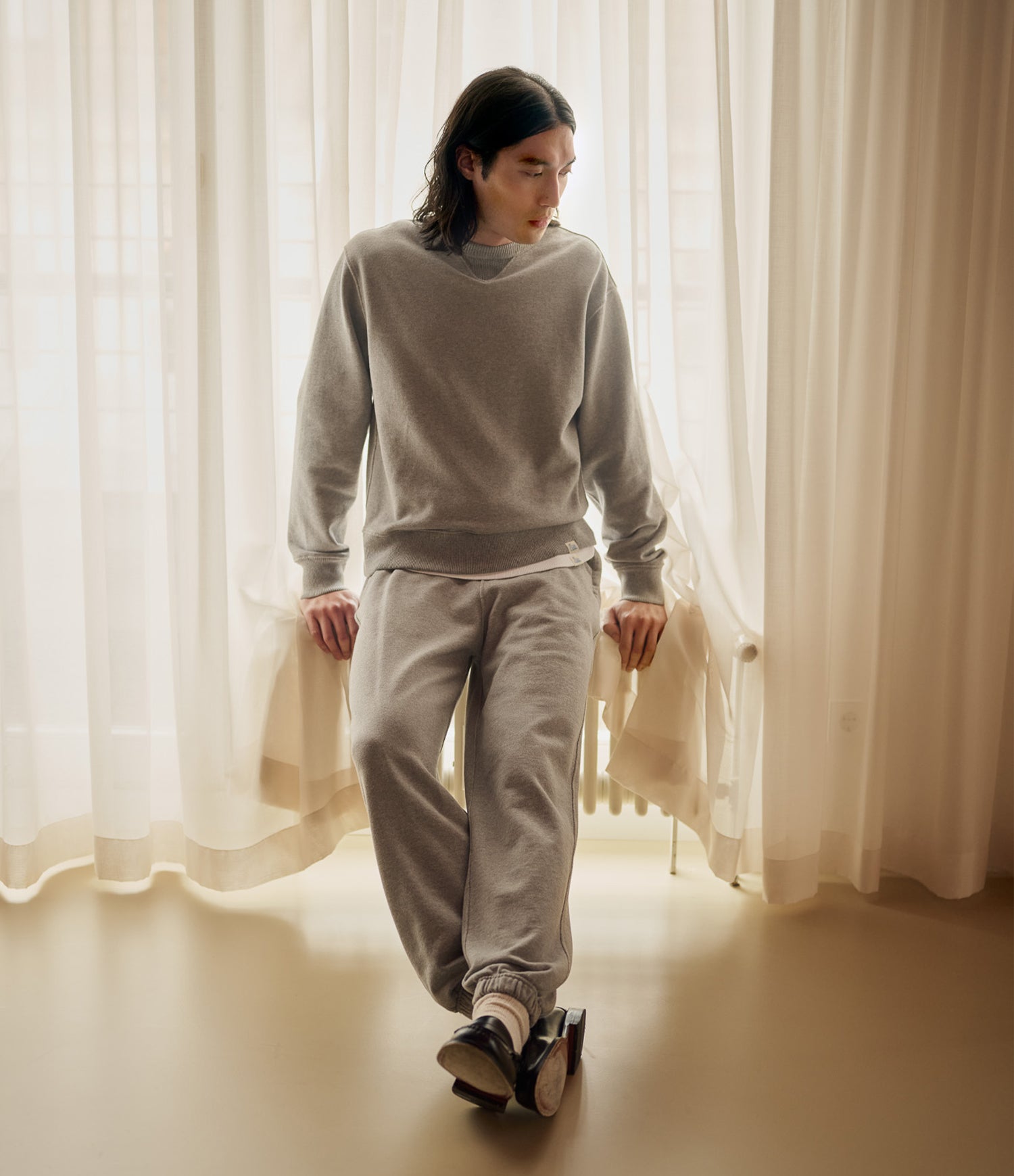 A person wearing a light gray crewneck sweatshirt and matching jogger sweatpants stands in a softly lit room with sheer white curtains and a beige floor.