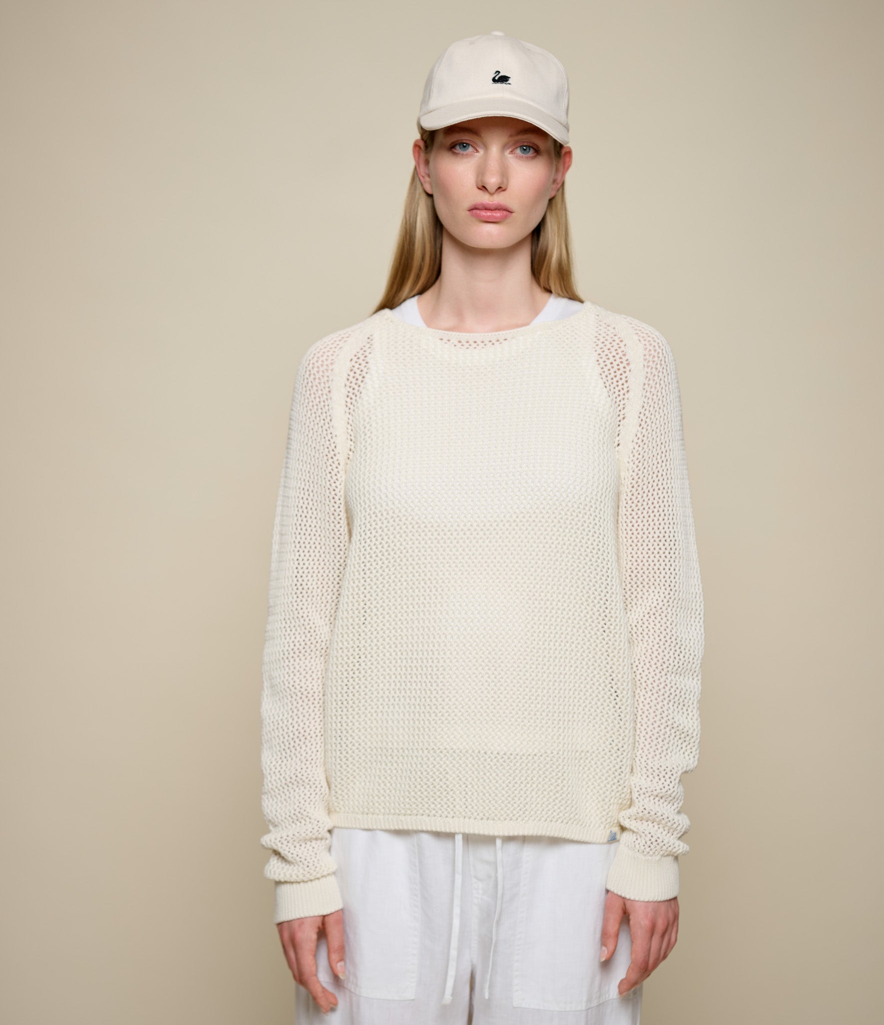 Woman wearing a beige knitted crochet sweater and cap against a beige background