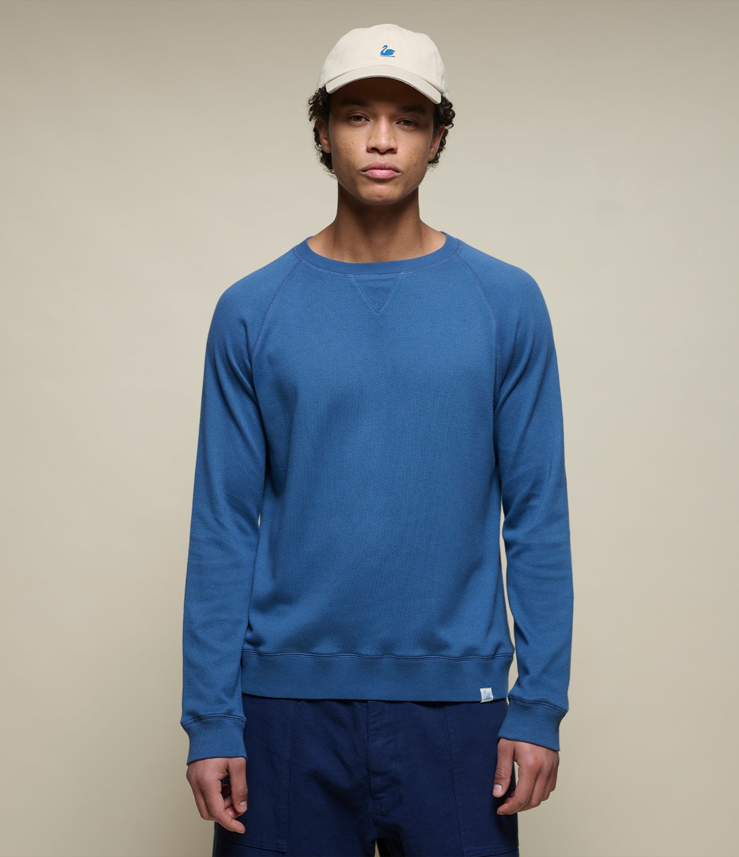 Person wearing an indigo blue lightweight sweatshirt and white cap on a beige background