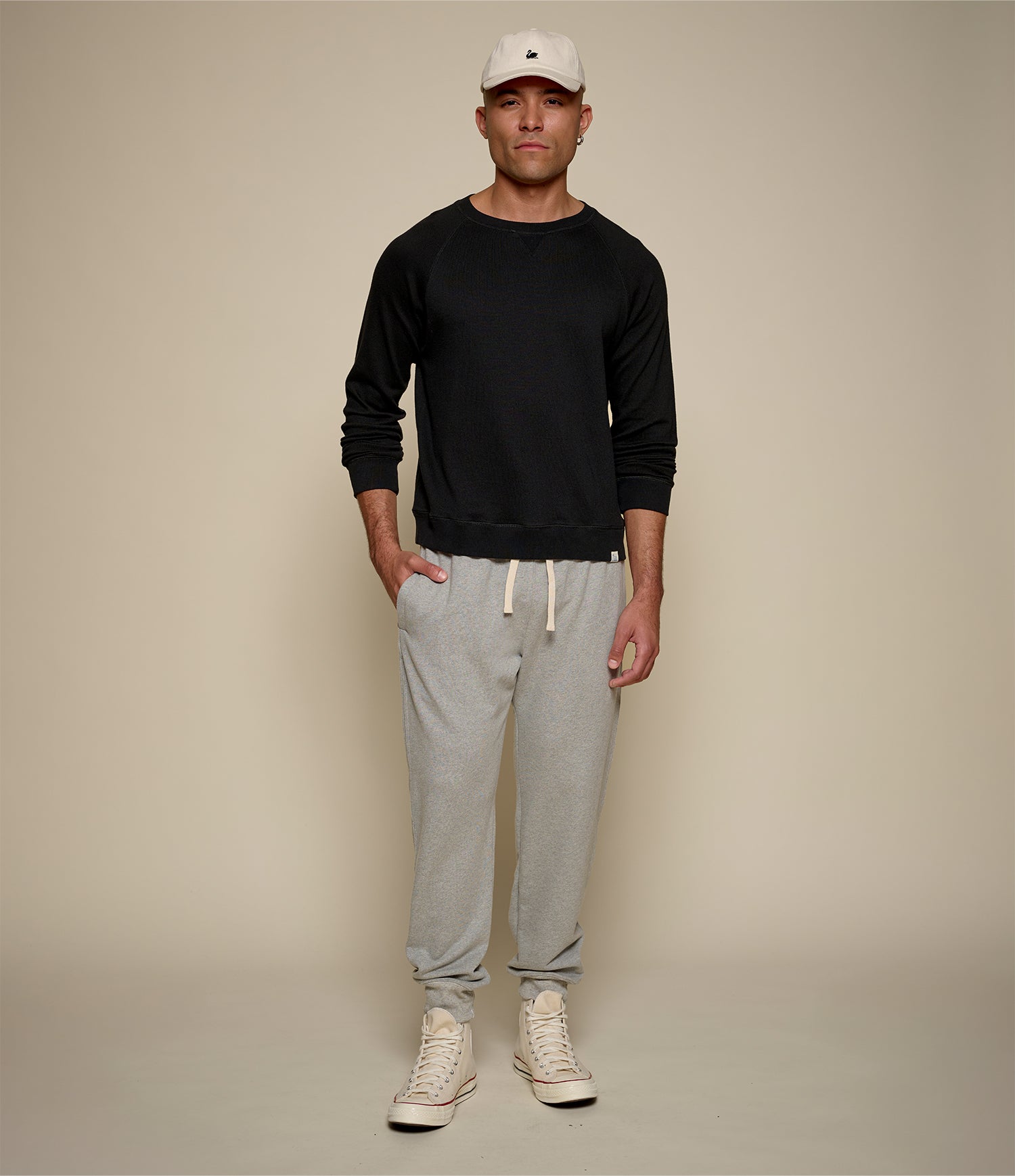 Man wearing a black long-sleeve shirt, gray sweatpants, and white sneakers on a beige background