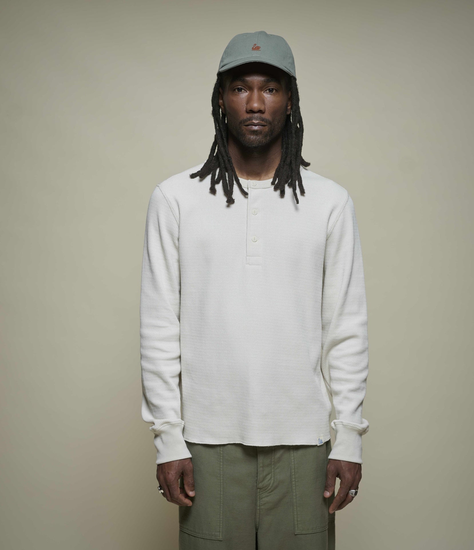 Man wearing a beige long-sleeve thermal henley shirt and green cap against a beige background