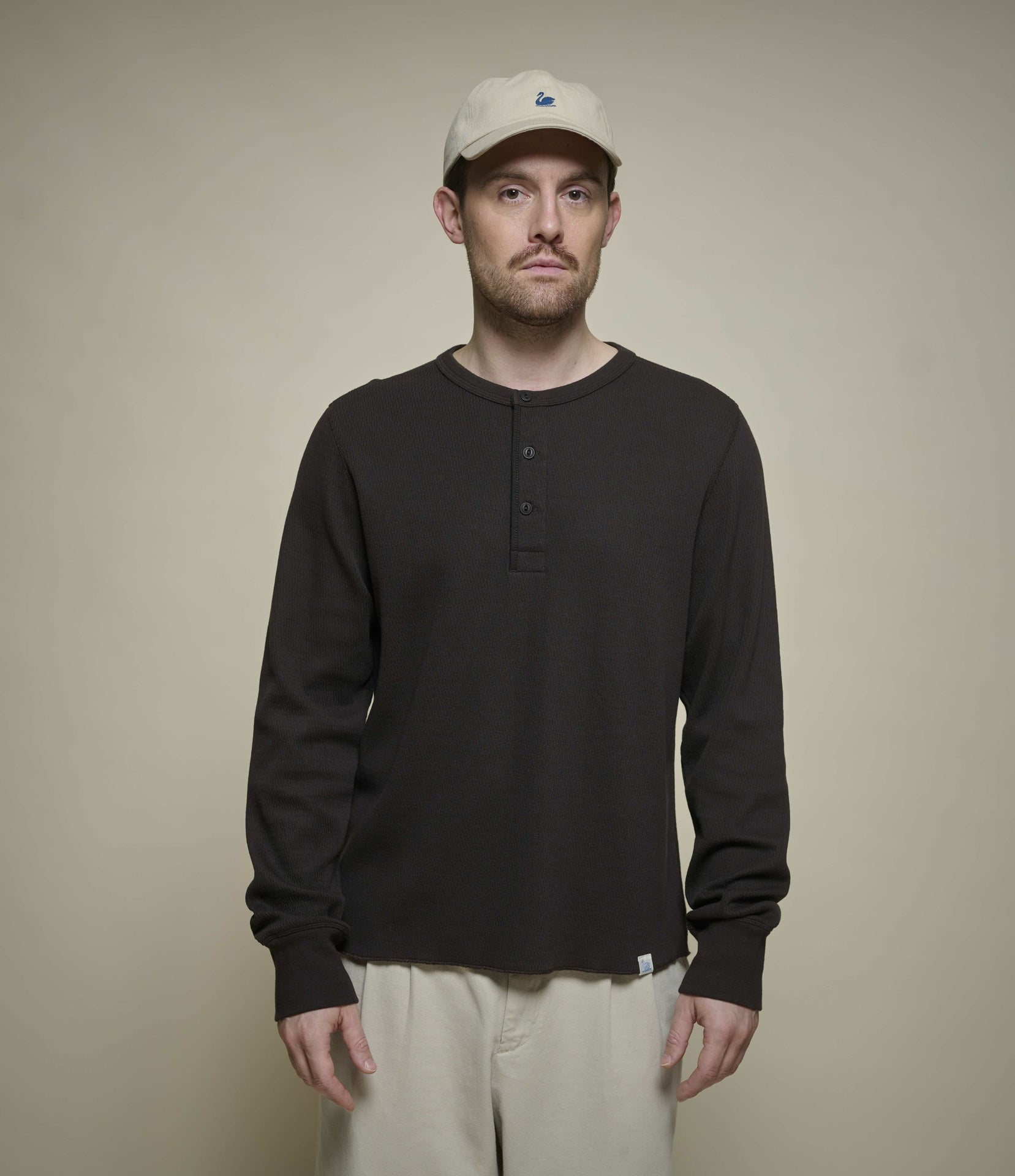 Man wearing a black long-sleeve thermal henley shirt and beige cap against a beige background