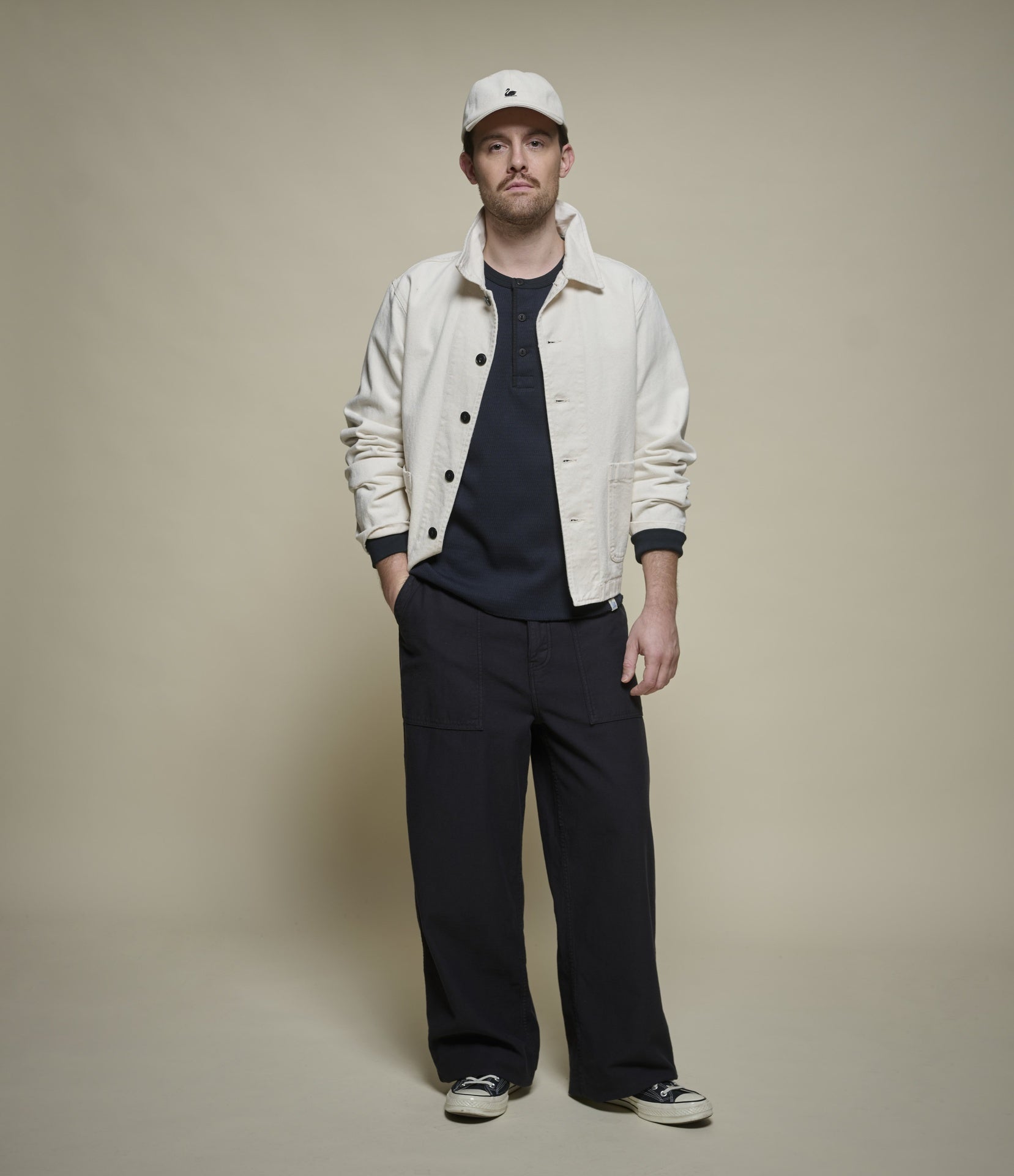 Man wearing a white jacket, black henley shirt, and black pants against a beige background