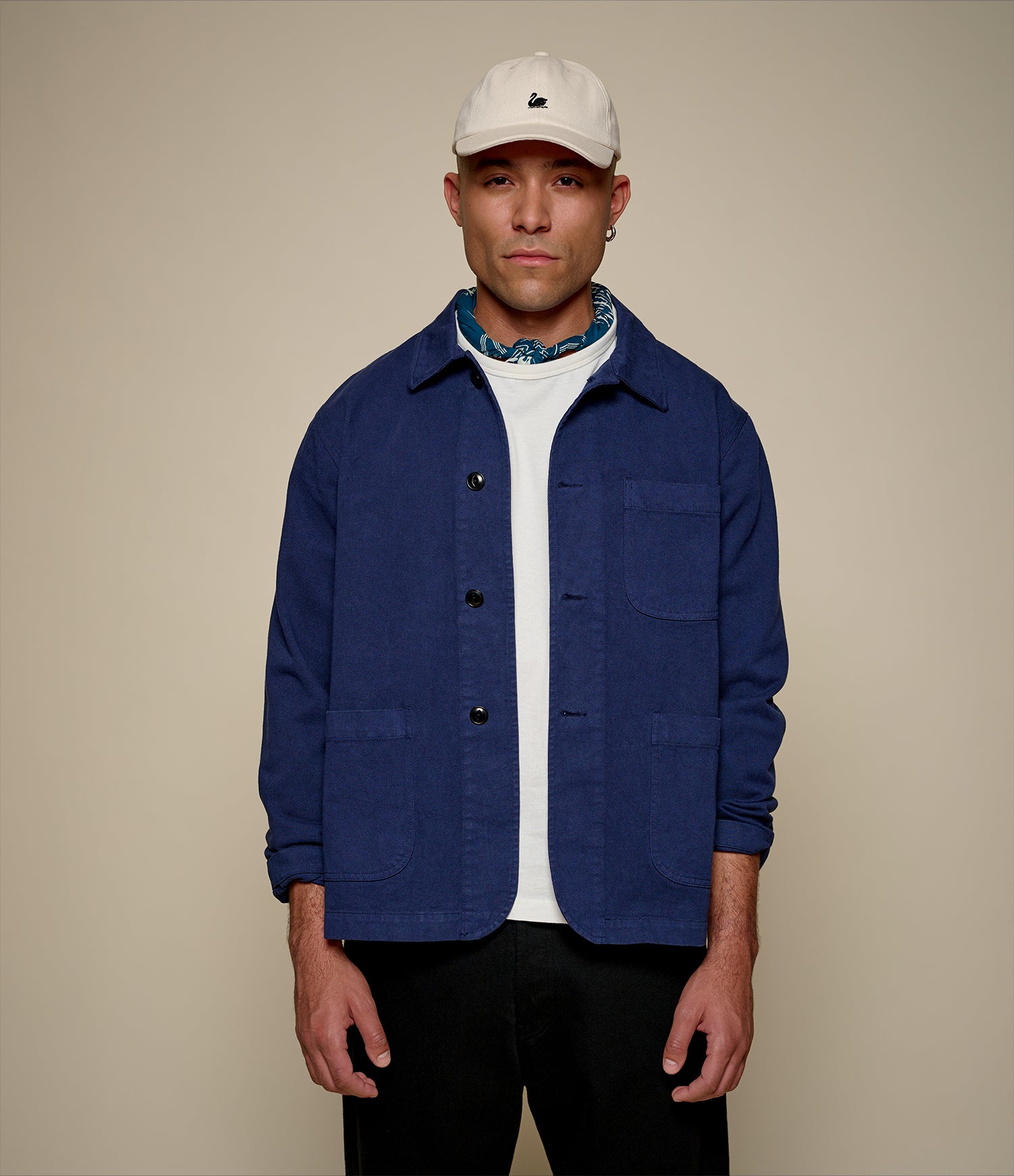 Man wearing a blue twill jacket, white shirt, and beige cap on a beige background