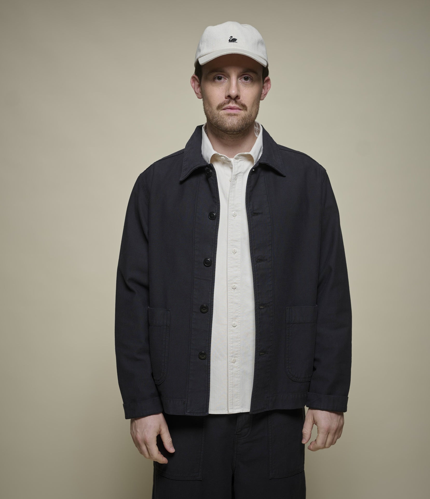 Man wearing a black cropped utility jacket over a beige shirt and with a white cap