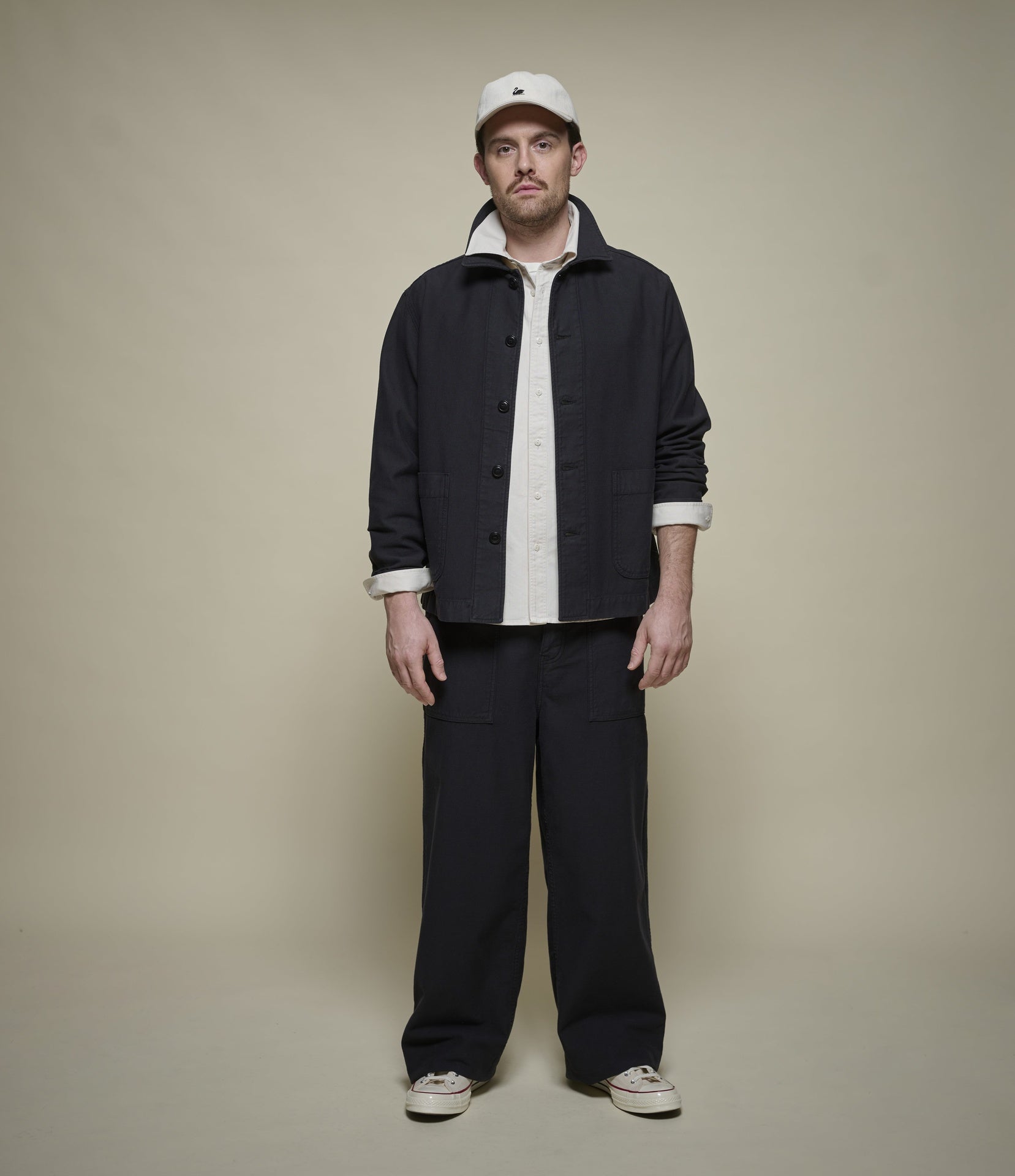 A man stands against a plain beige studio background wearing a dark black utility jacket over a white button-up shirt, matching dark navy trousers, and a white baseball cap.