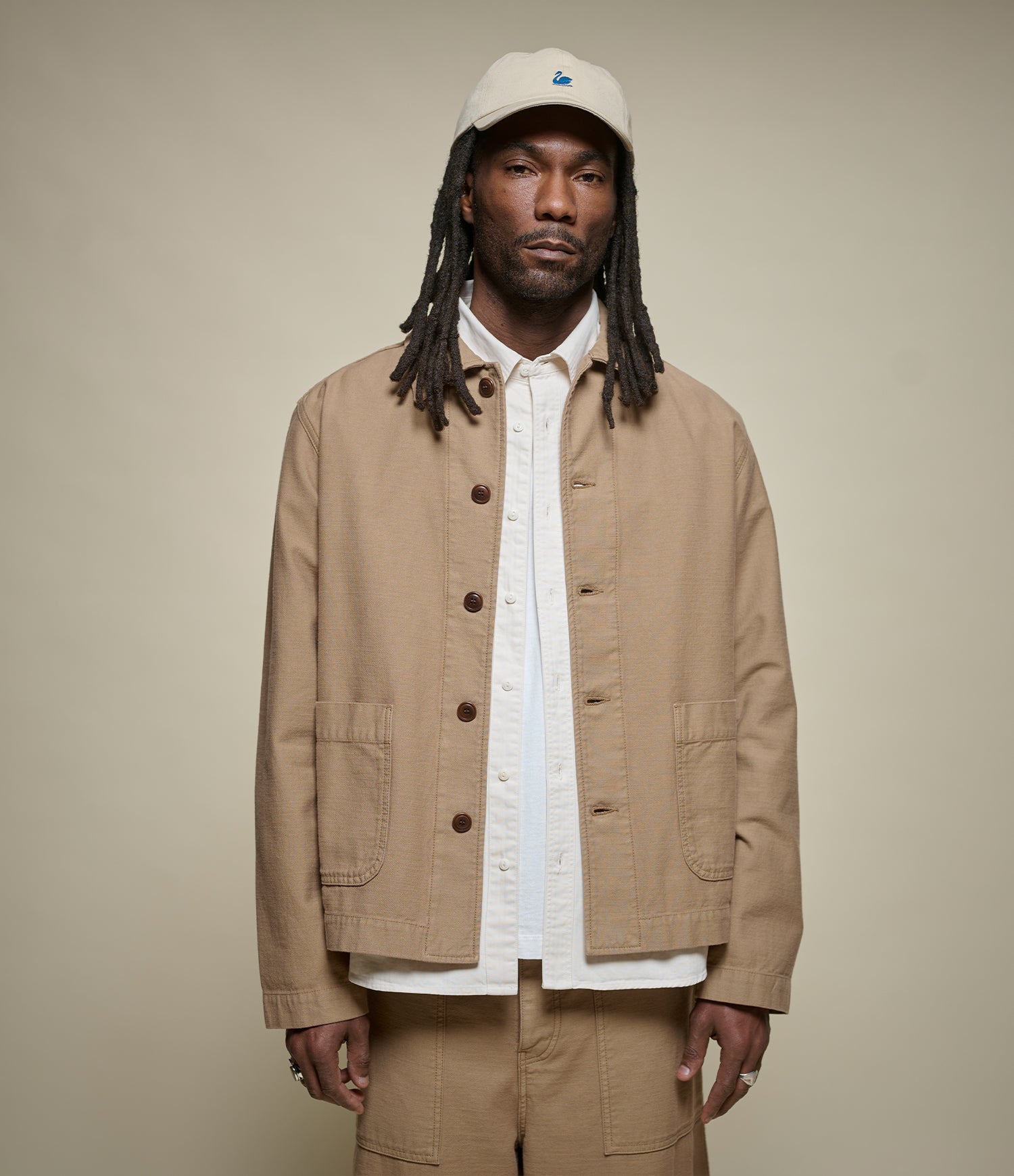 A man wearing a beige utility jacket over a white button-up shirt, matching beige pants, and a beige baseball cap, standing against a solid beige background.