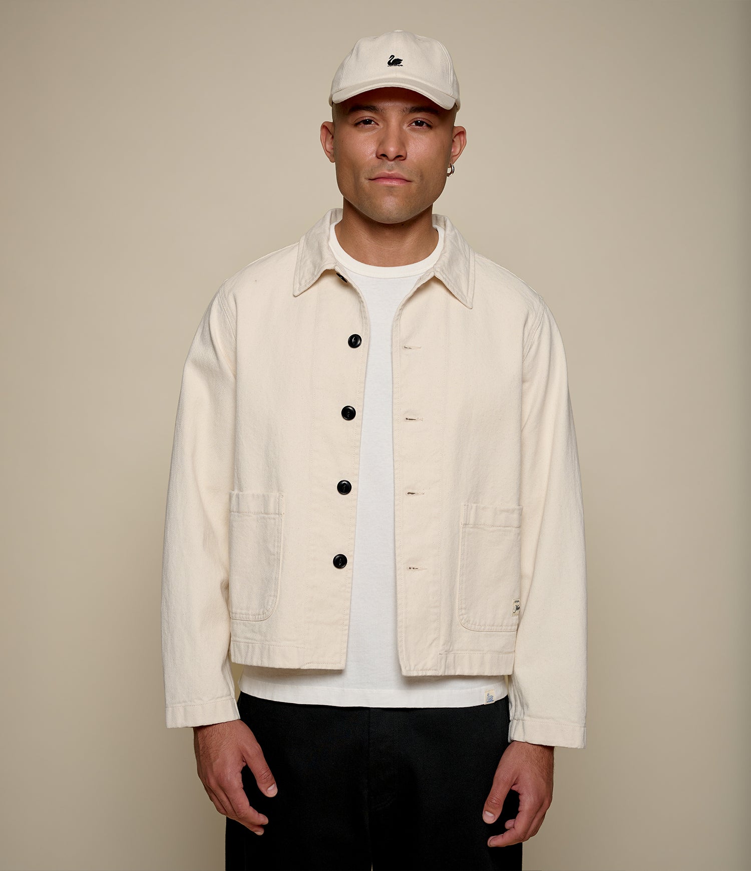 Man wearing a beige cropped twill jacket, white shirt, and cap against a beige background