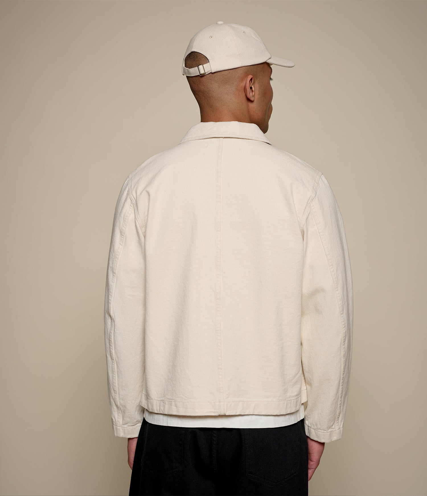 Man wearing a beige cropped twill jacket and cap from behind on a beige background