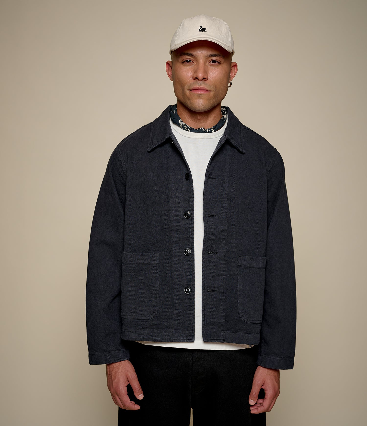 Man wearing a dark cropped twill jacket, white shirt, and cap against a beige background