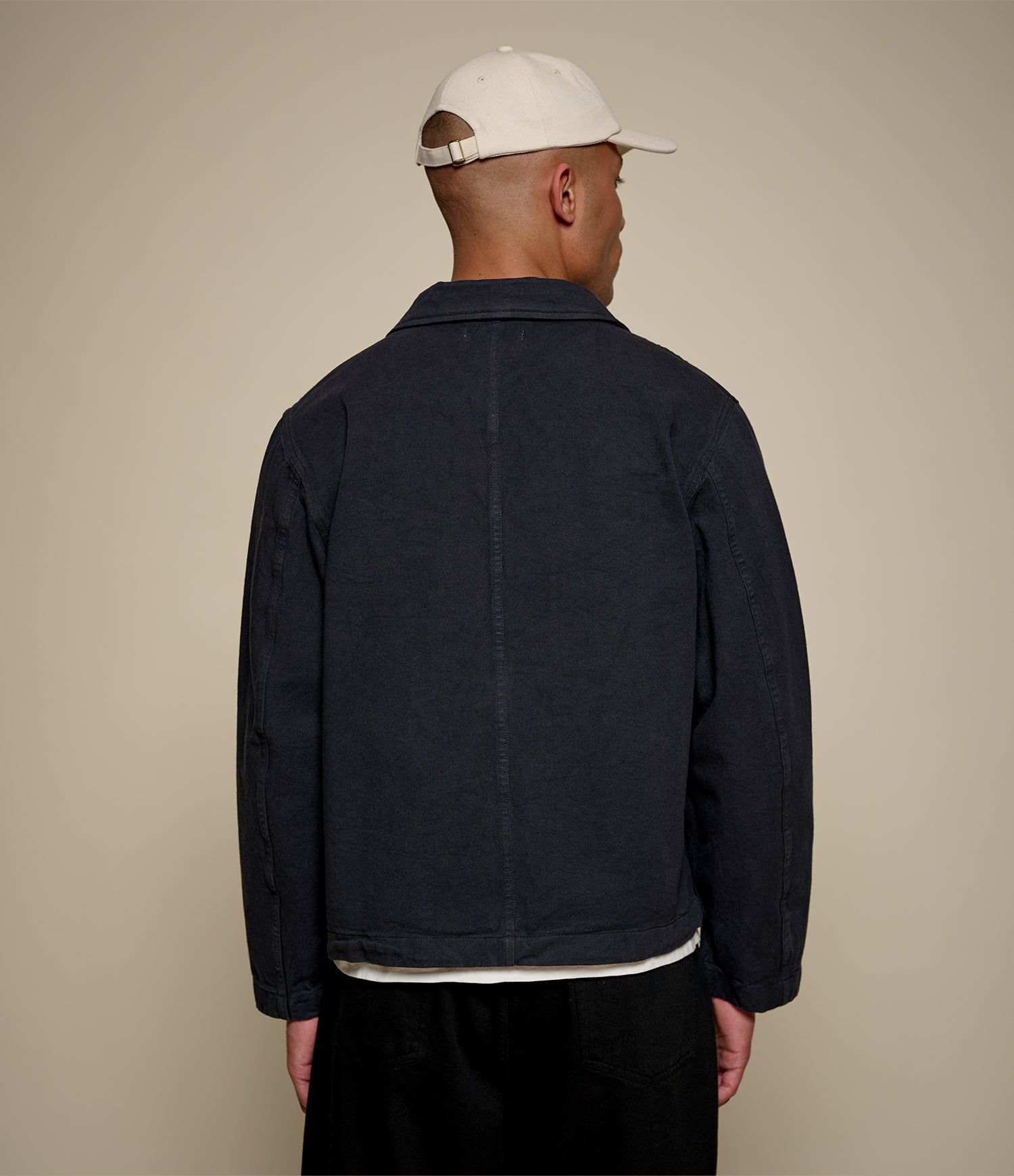 Man wearing a black cropped twill jacket and white cap on a beige background