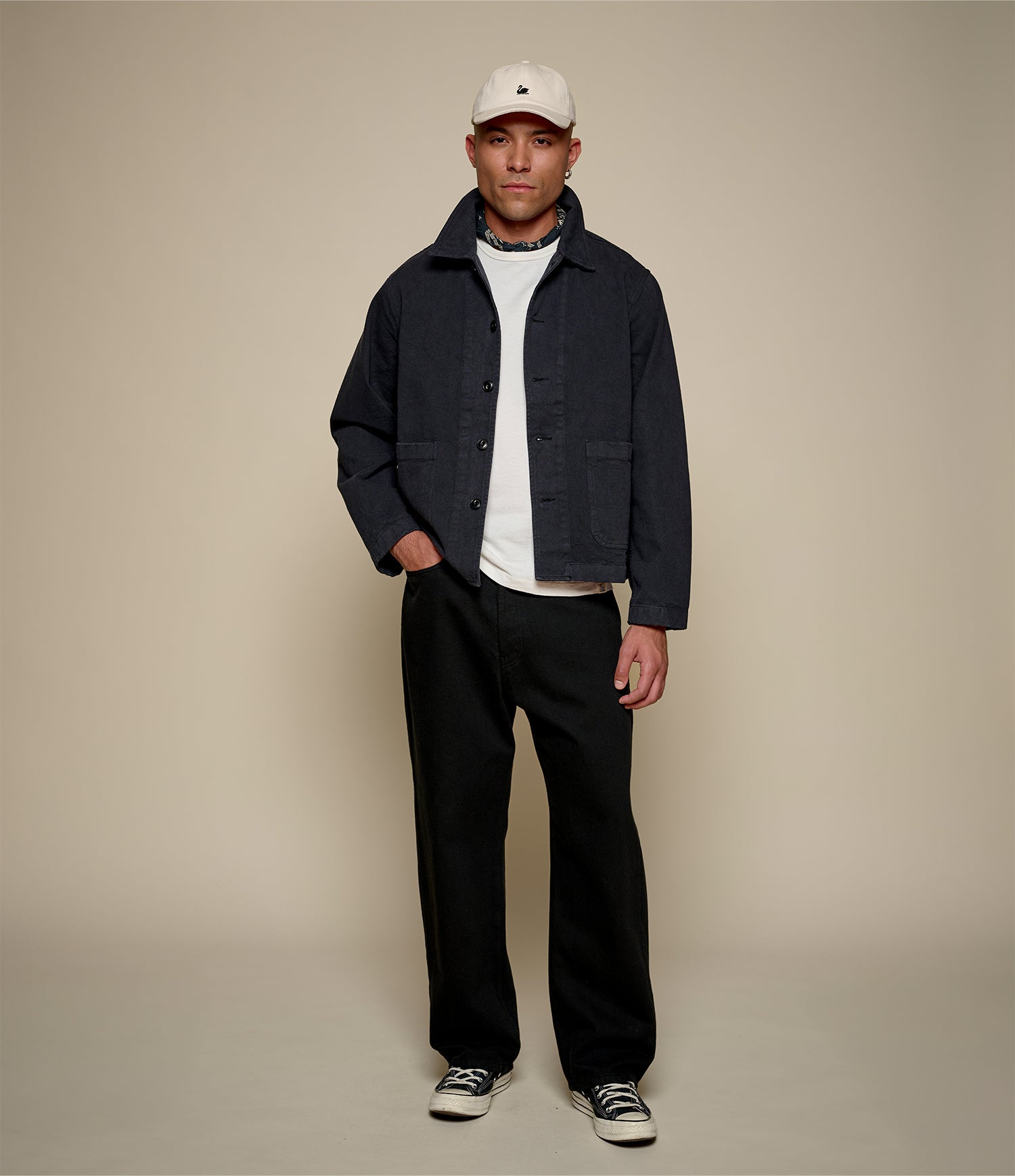 Man wearing a dark cropped twill jacket, white shirt, black pants, and a cap on a beige background