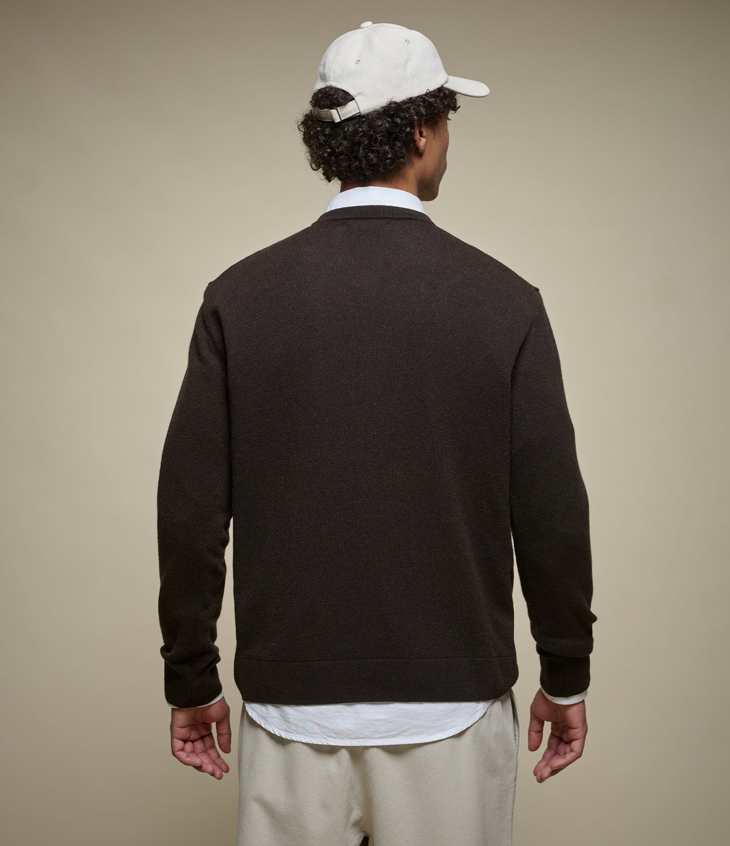 A man shown from the back wearing a dark brown knit henley sweater over a white collared shirt, paired with light beige trousers and a white baseball cap, against a solid beige studio background.