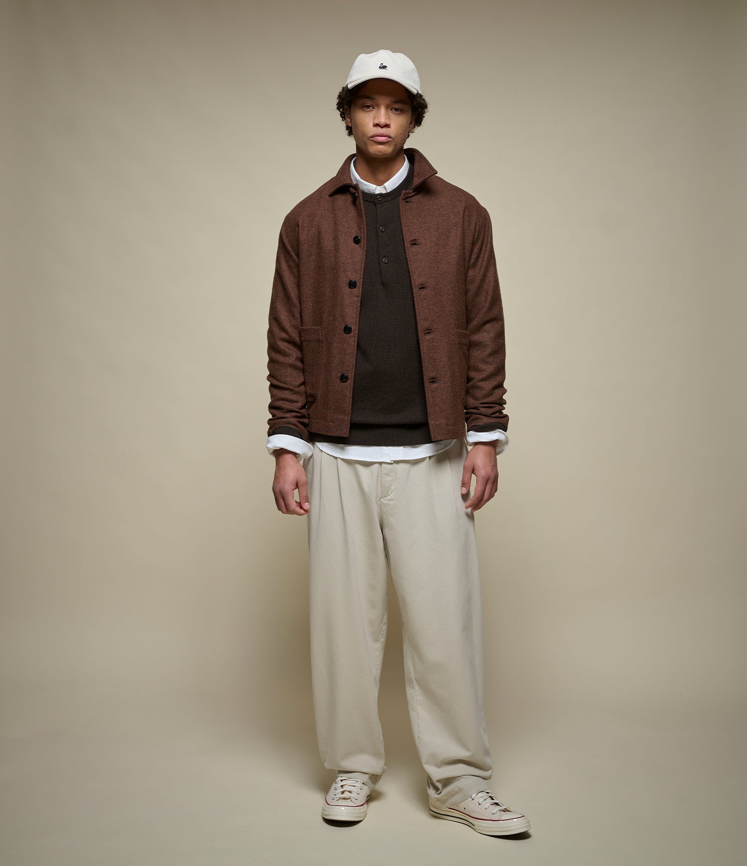 A model stands against a solid beige studio background wearing a brown textured button-up jacket over a dark henley sweater and white shirt, paired with beige trousers and a white cap.