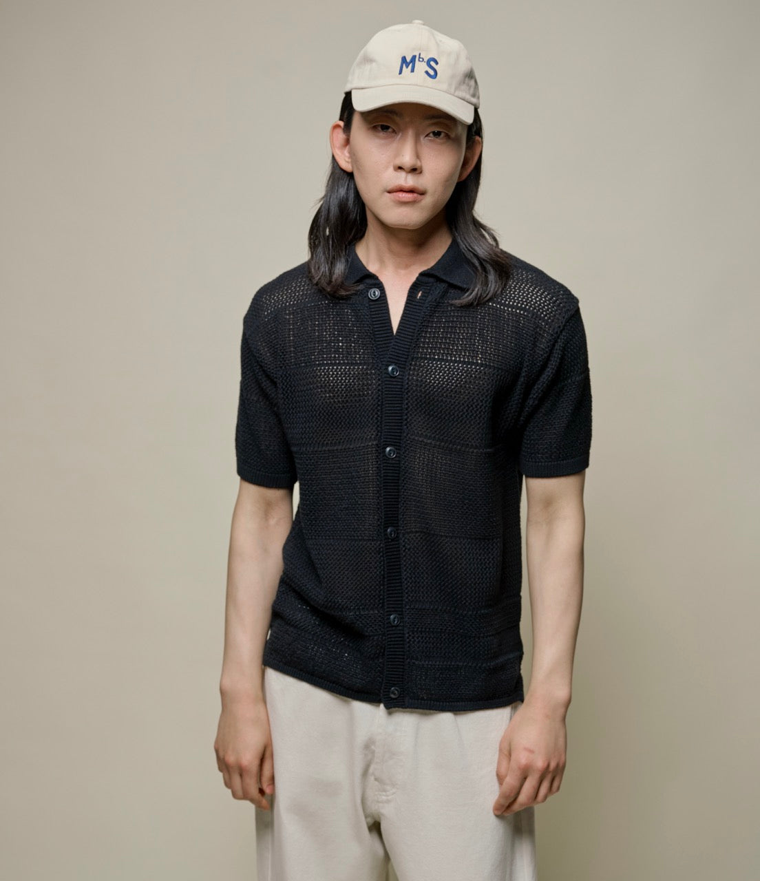 A person wearing a black short sleeve crochet cotton shirt with a ribbed collar and corozo nut buttons, paired with light-colored pants and a white cap with the letters 'MS' on it.