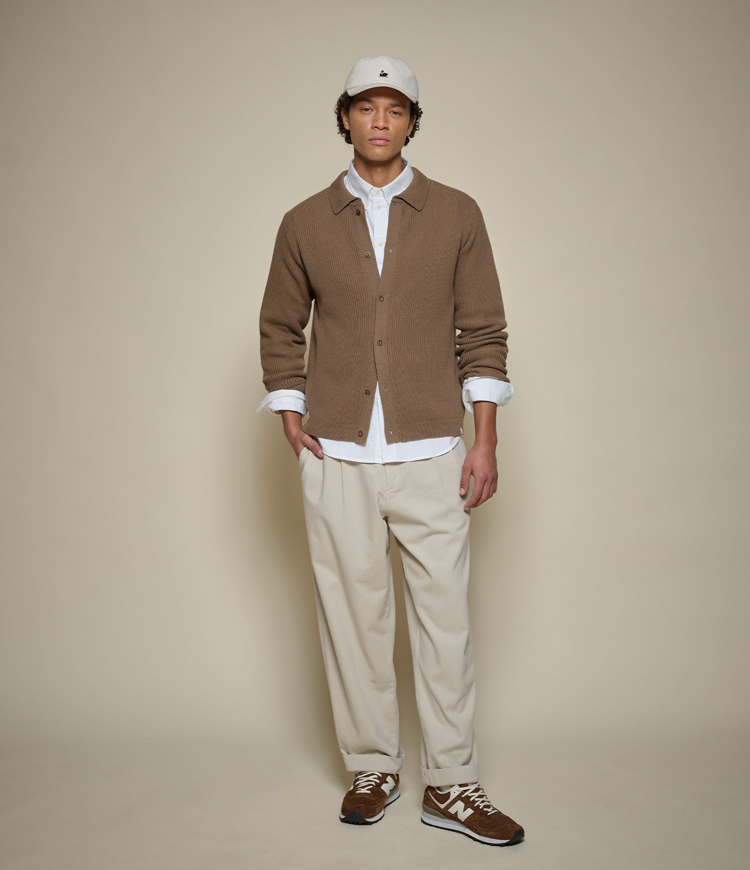 A man modeling a brown button-up cardigan over a white collared shirt, paired with beige trousers, brown sneakers, and a white baseball cap, against a solid beige studio background.