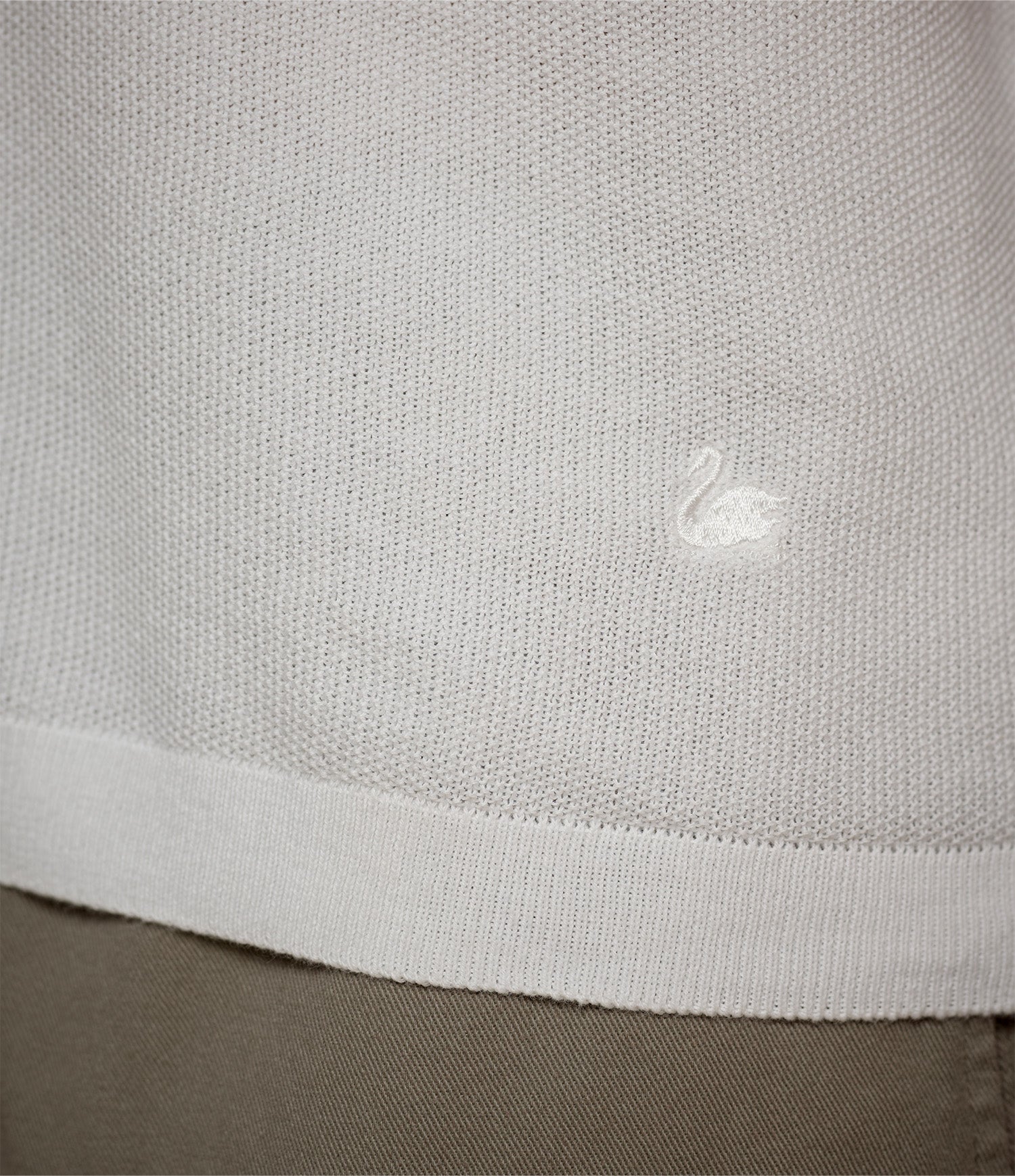 Close-up of a white knitted polo shirt with a small embroidered swan logo above the hem