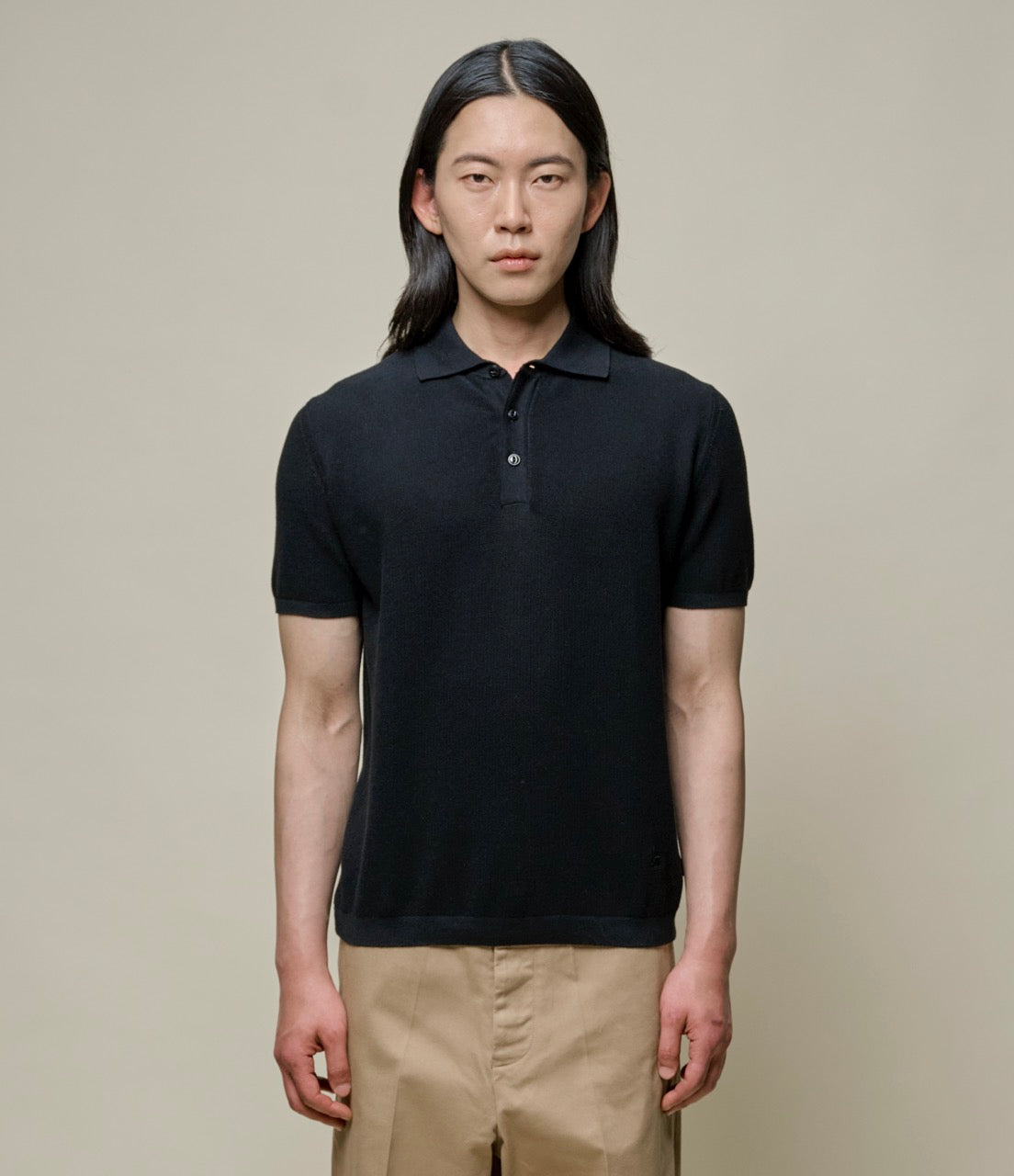 A person wearing a black knitted polo shirt with short sleeves, collar, and front buttons, paired with beige pants.