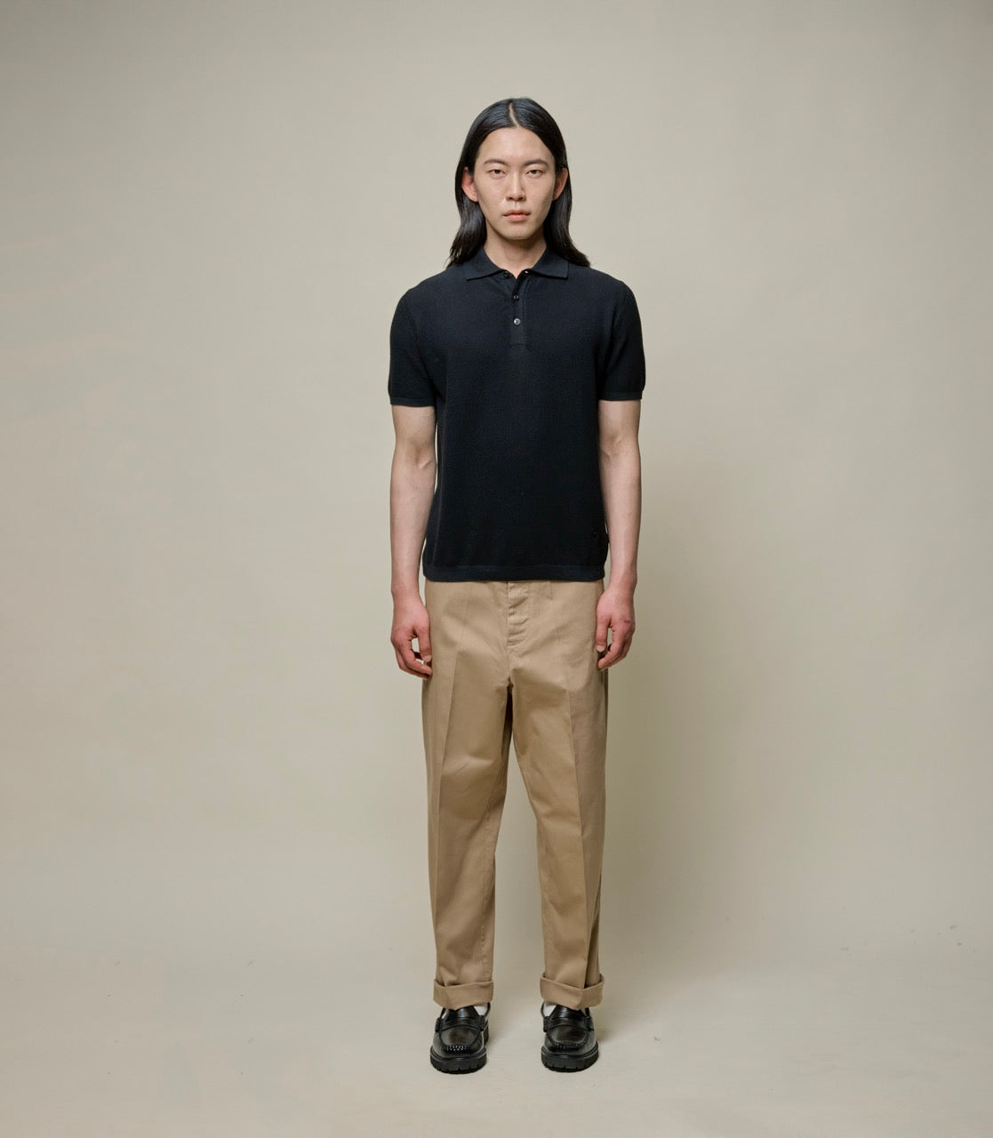 Person wearing a black knitted polo shirt and beige pants against a plain background