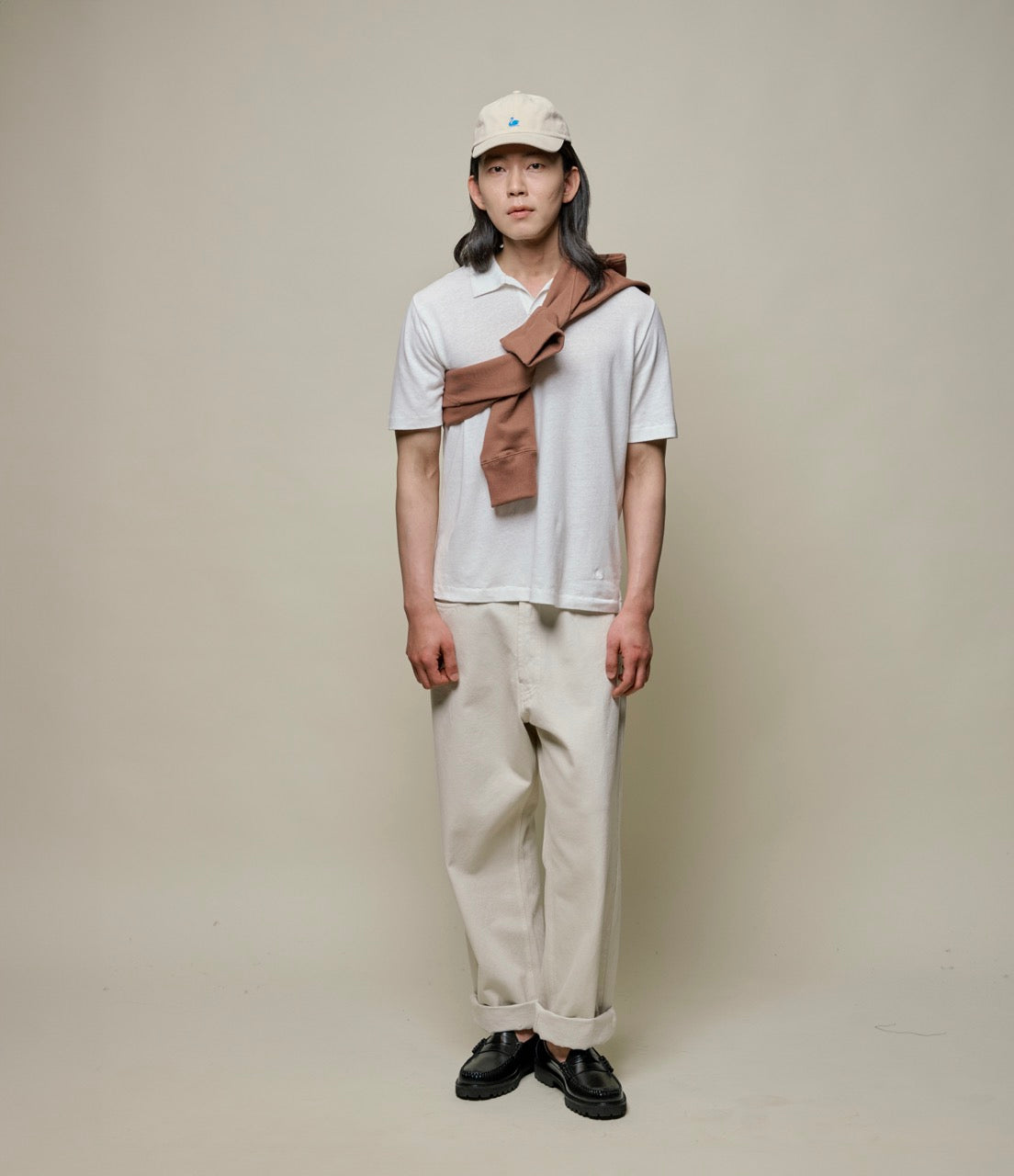 Person wearing a white linen polo shirt, beige pants, and a brown bag over one shoulder against a plain background