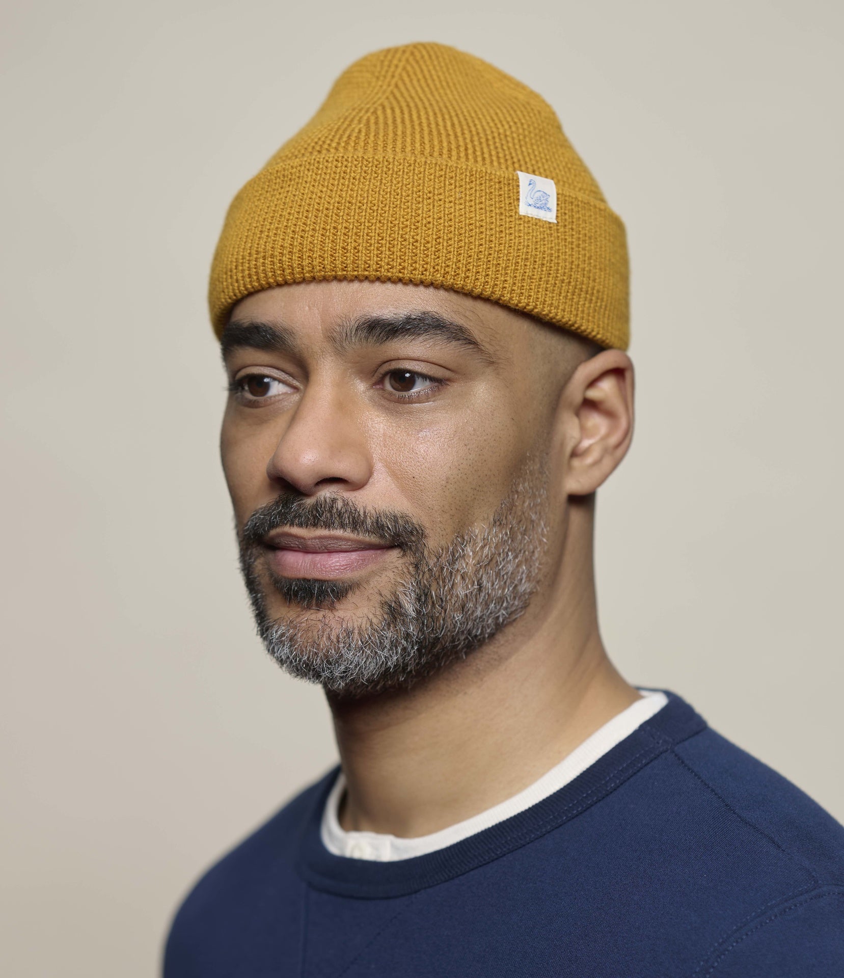 A man wearing a mustard yellow ribbed knit beanie hat with a small white brand tag, paired with a navy blue crewneck sweater. The background is a solid, warm off-white studio wall.