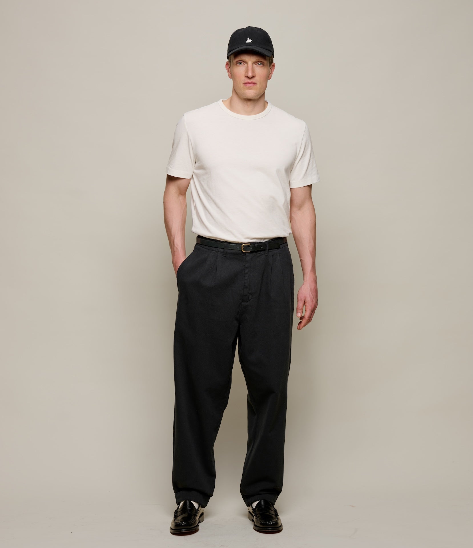 Man wearing a white t-shirt, black pleated pants, and a black cap on a plain background