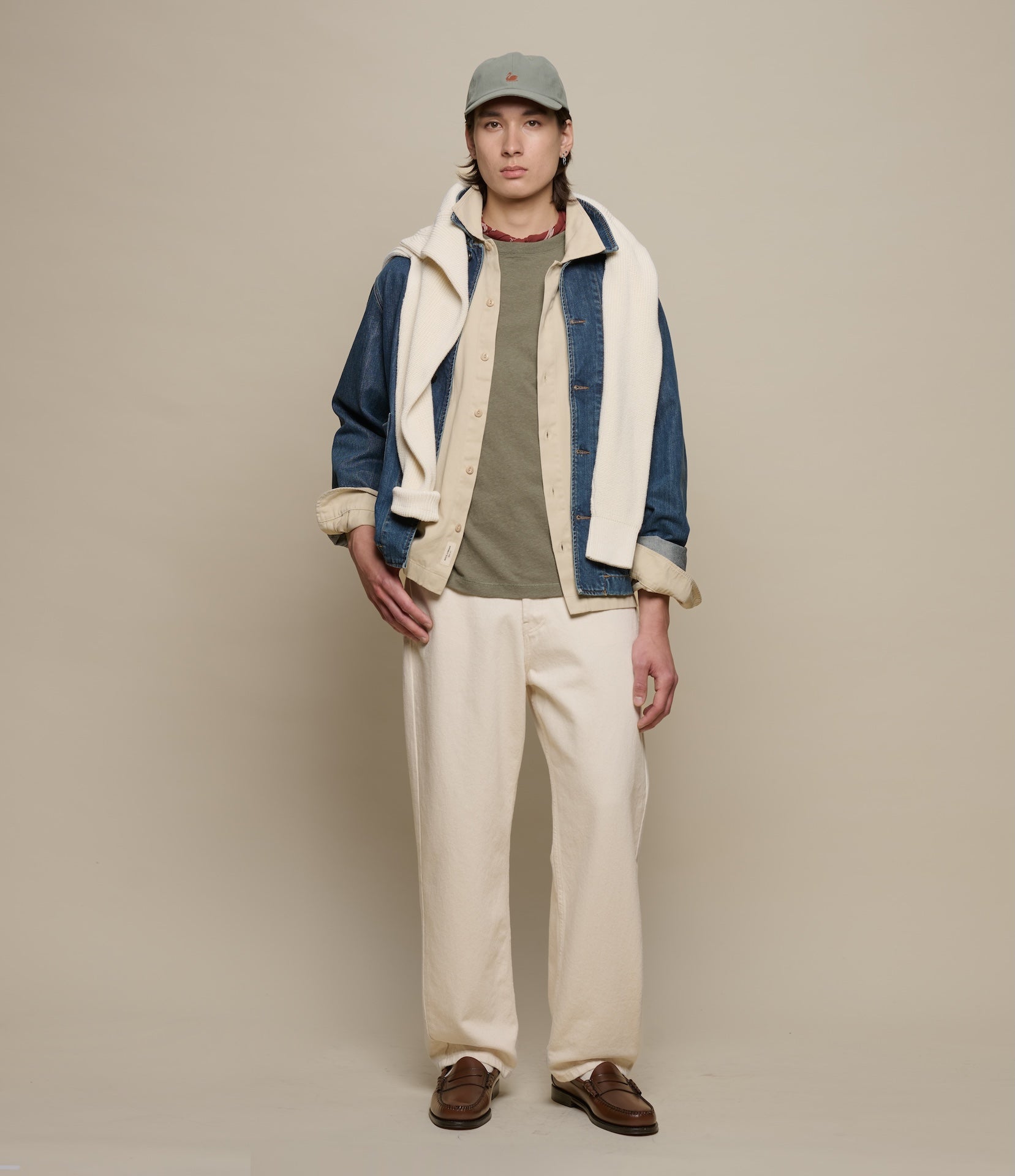 Person wearing a layered outfit with a jacket, shirt, pants, and cap on a plain background