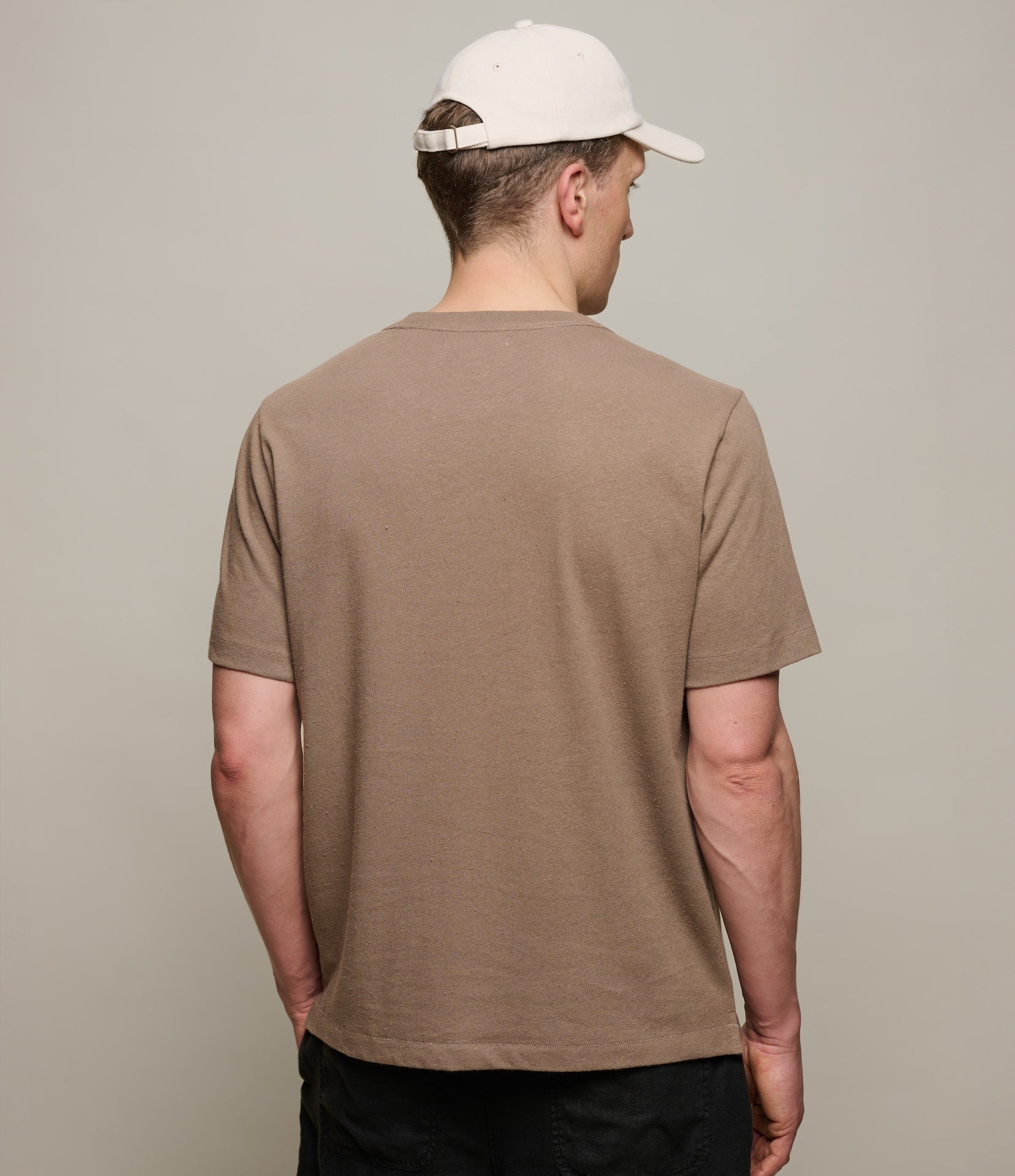 Man wearing a brown linen t-shirt and beige cap from behind on a plain background
