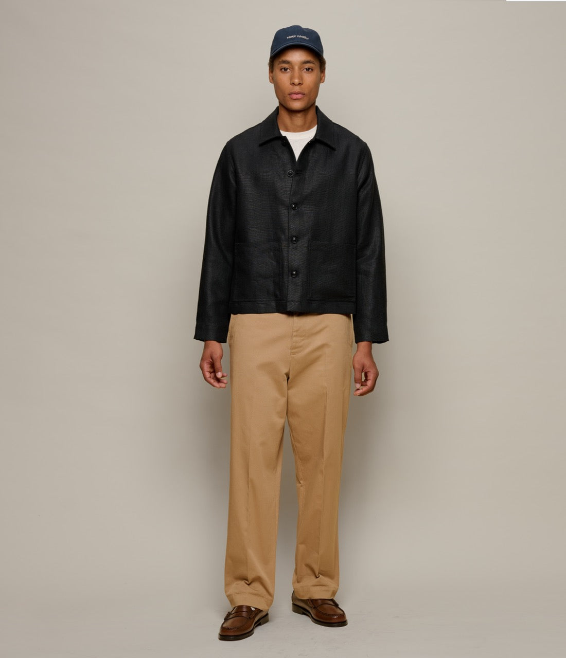 Man wearing a black linen jacket, beige pants, and a blue cap on a plain background