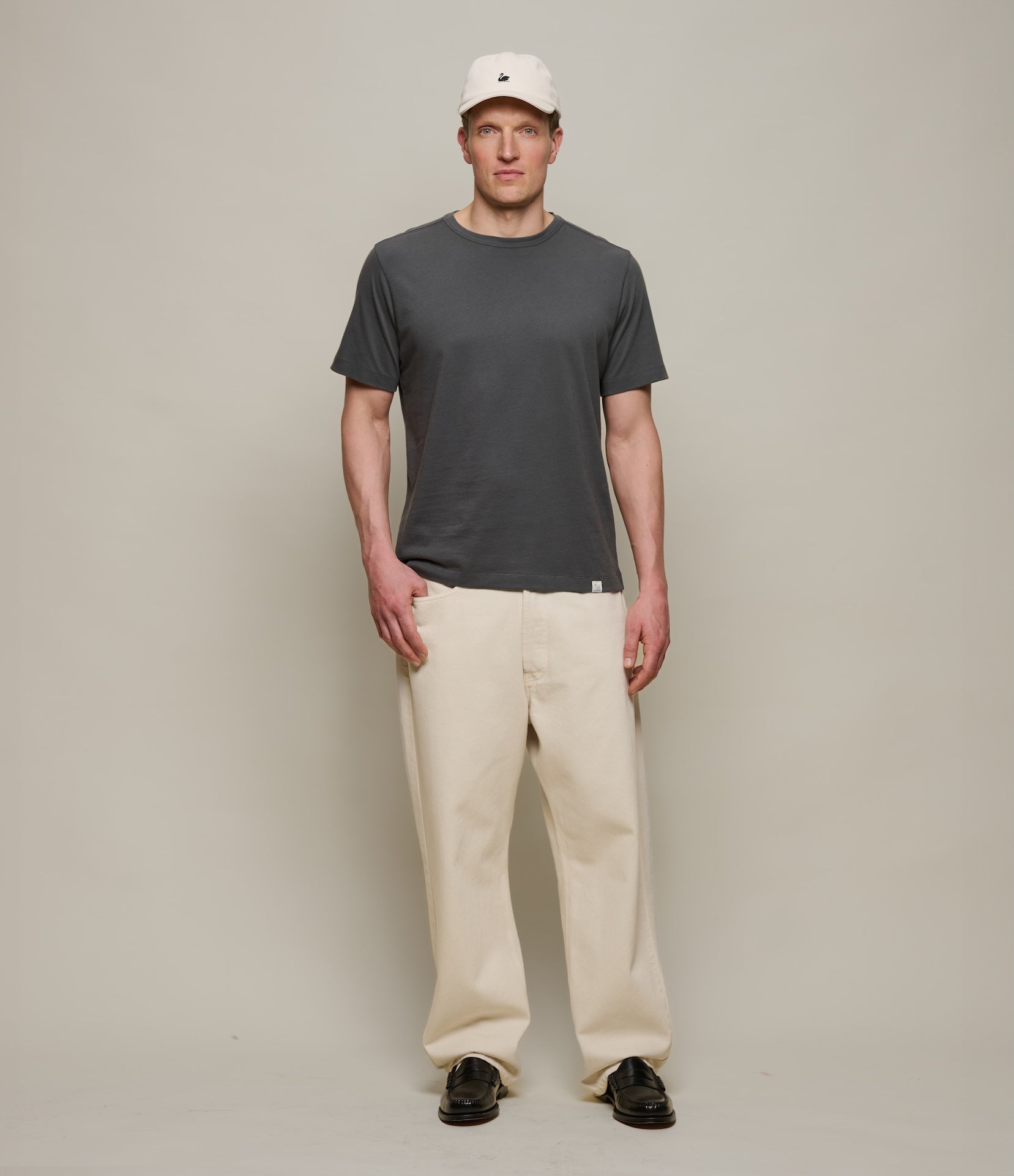 Man wearing a dark gray t-shirt, beige pants, and a white cap on a plain background