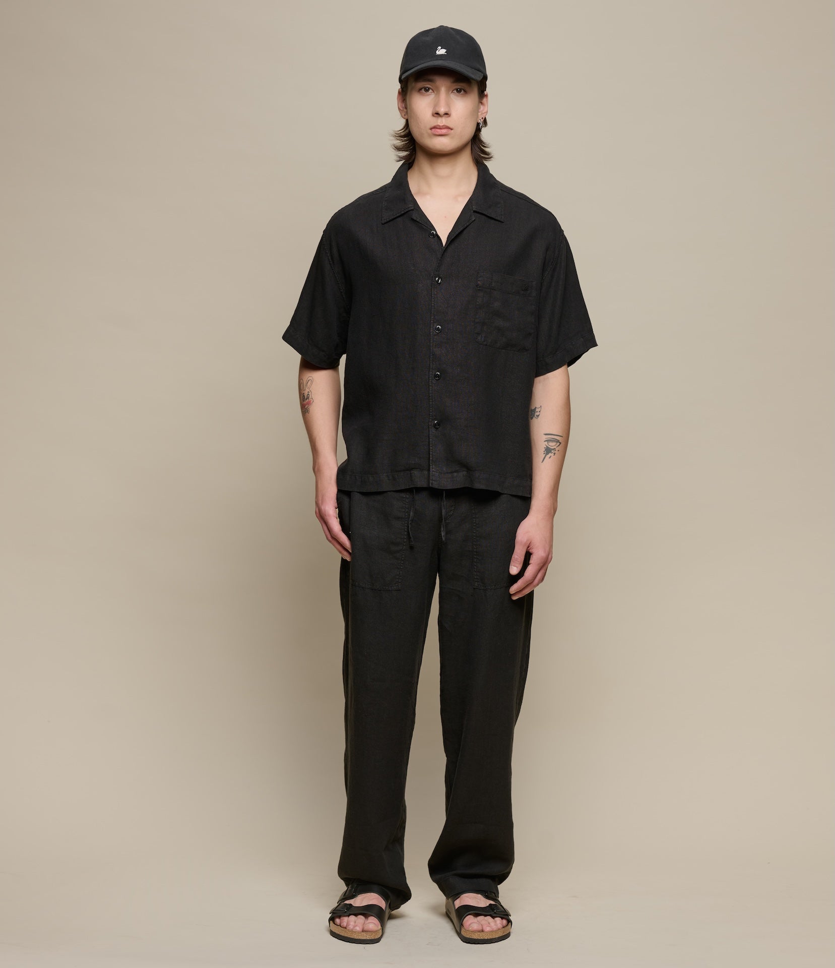 Person wearing a black linen outfit with a cap on a beige background