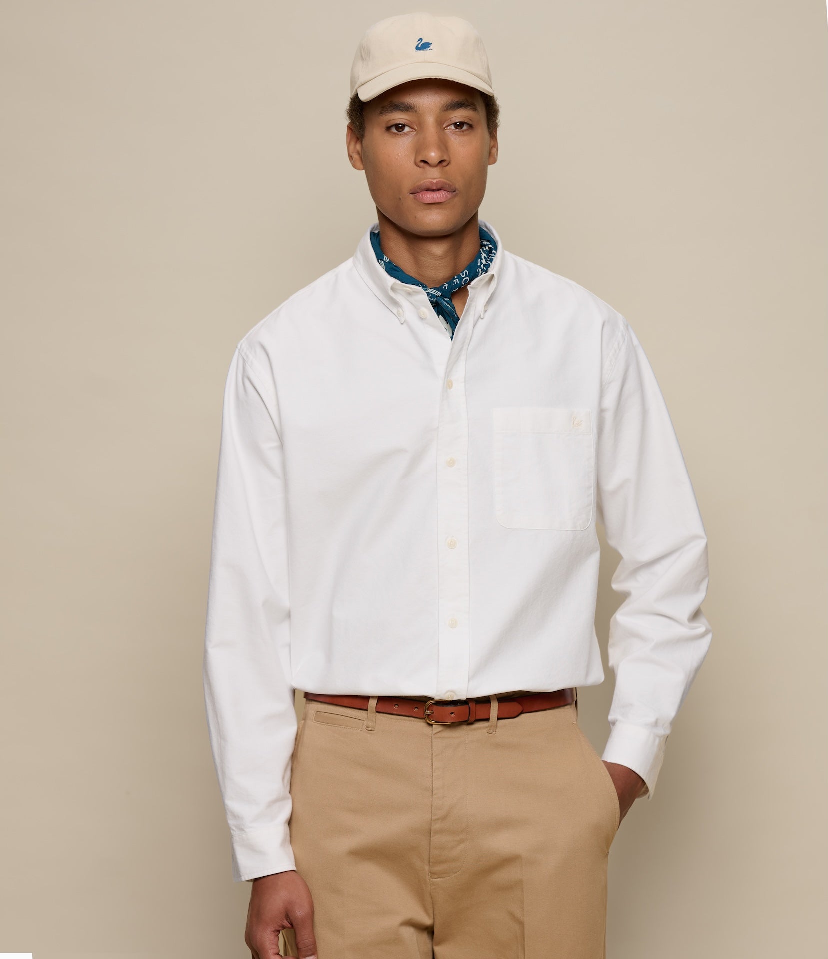 Man wearing a white button-down shirt, beige cap, and khaki pants against a beige background