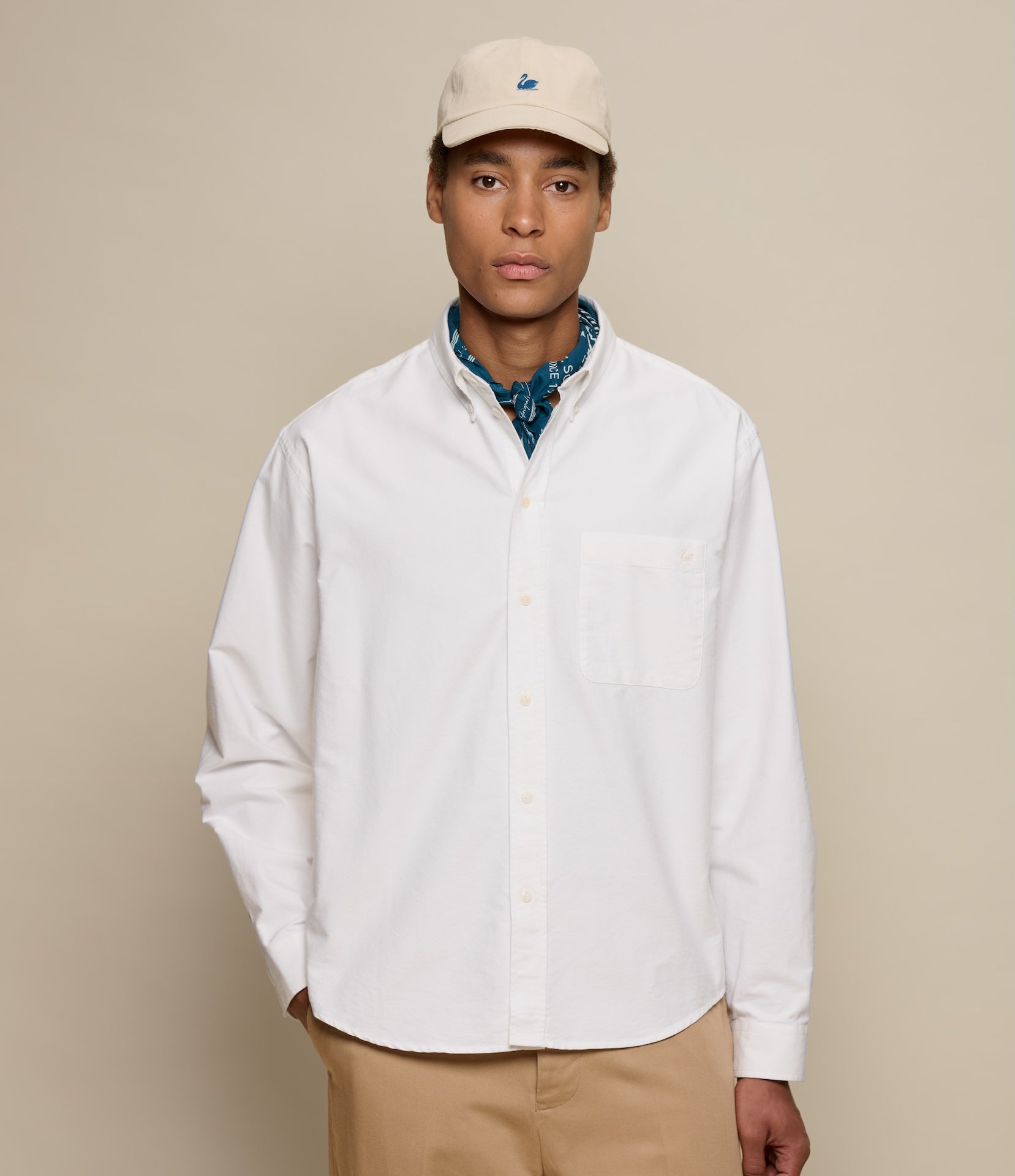 Person wearing a white button-down shirt, beige cap, and beige pants against a beige background