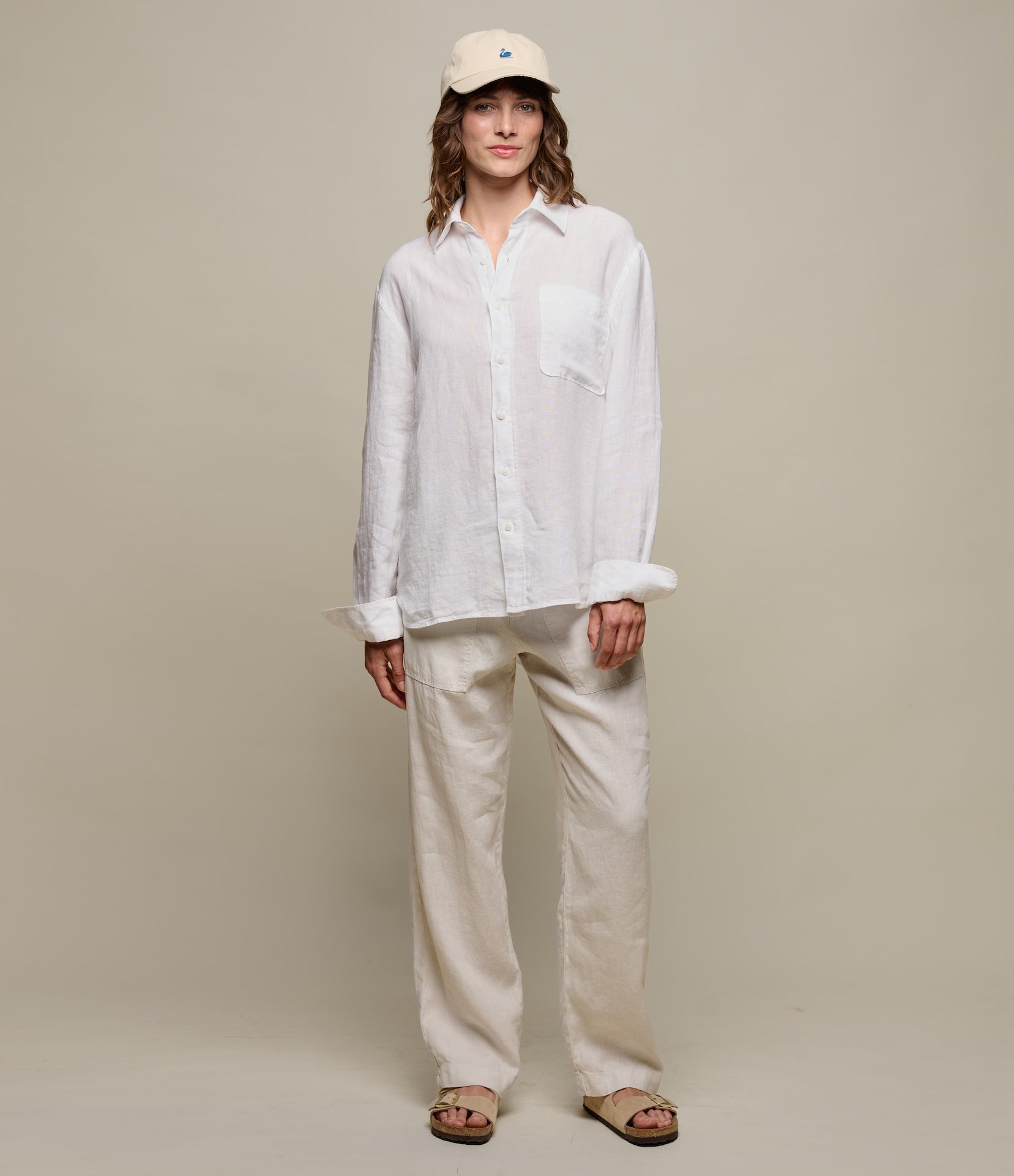 Person wearing a white linen shirt and pants with a beige cap on a plain background