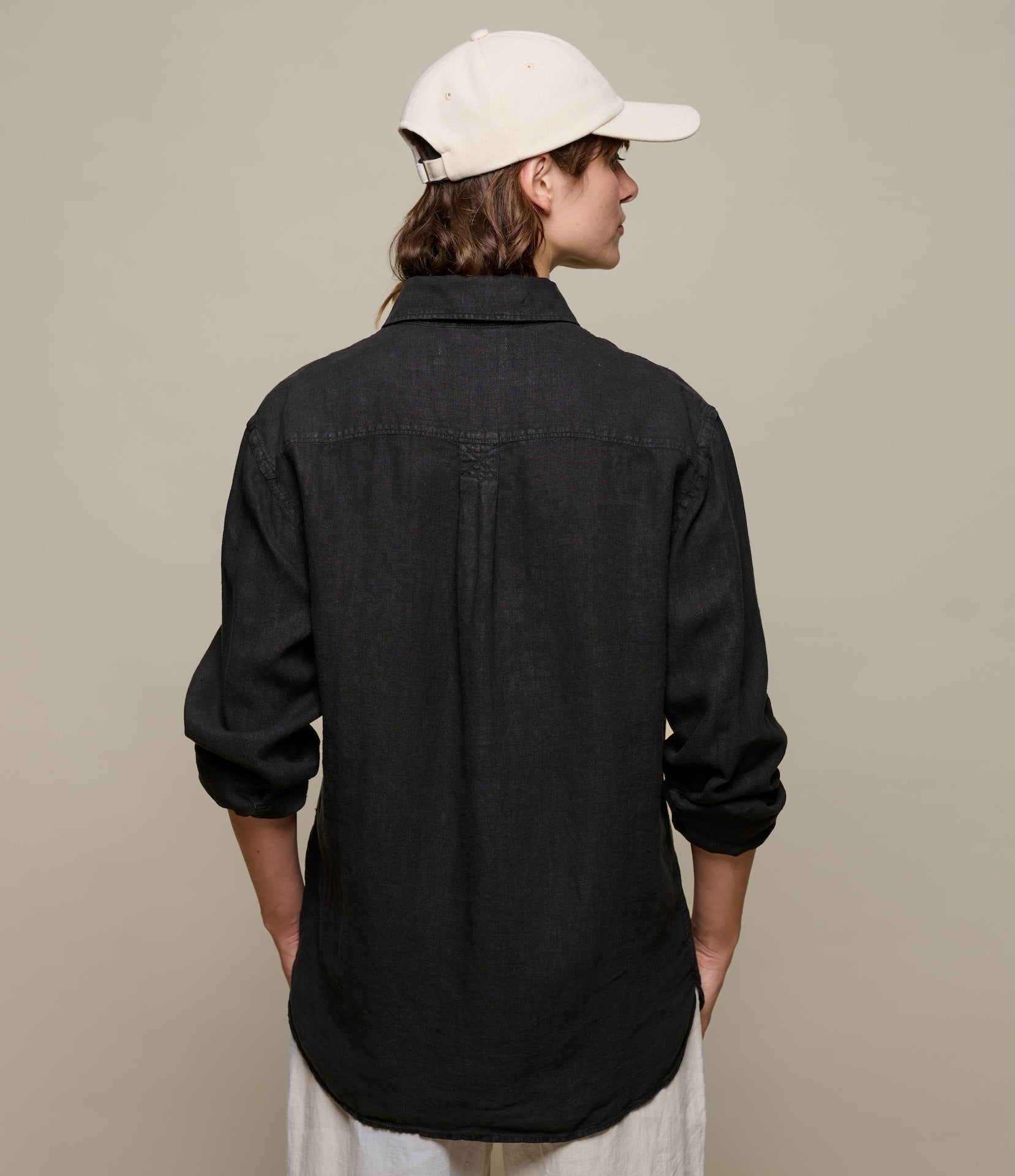 Person wearing a black linen shirt and white cap from behind on a beige background